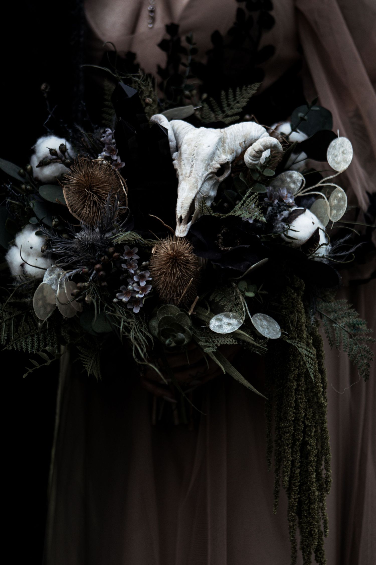 Dark gothic wedding bouquet with animal skull, cotton flowers, dried botanicals, and dark foliage held by person in gray gown.