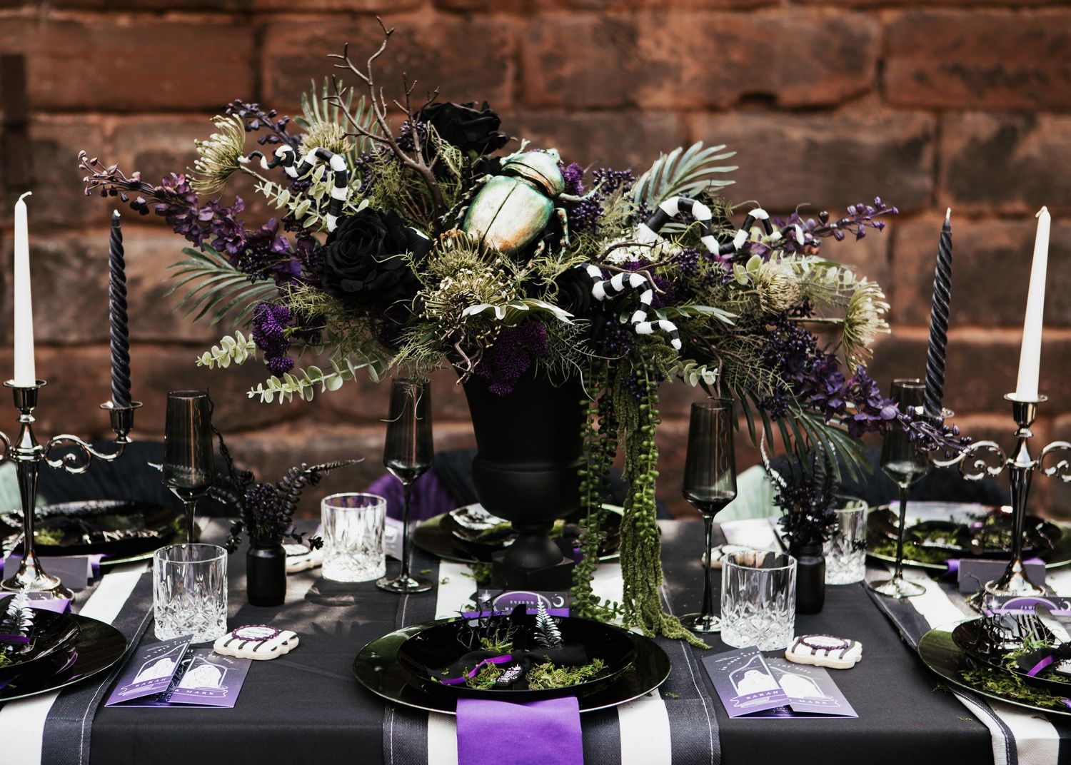 Gothic wedding table with black and purple floral centerpiece, dark plates, candles, and elegant purple ribbon accents.