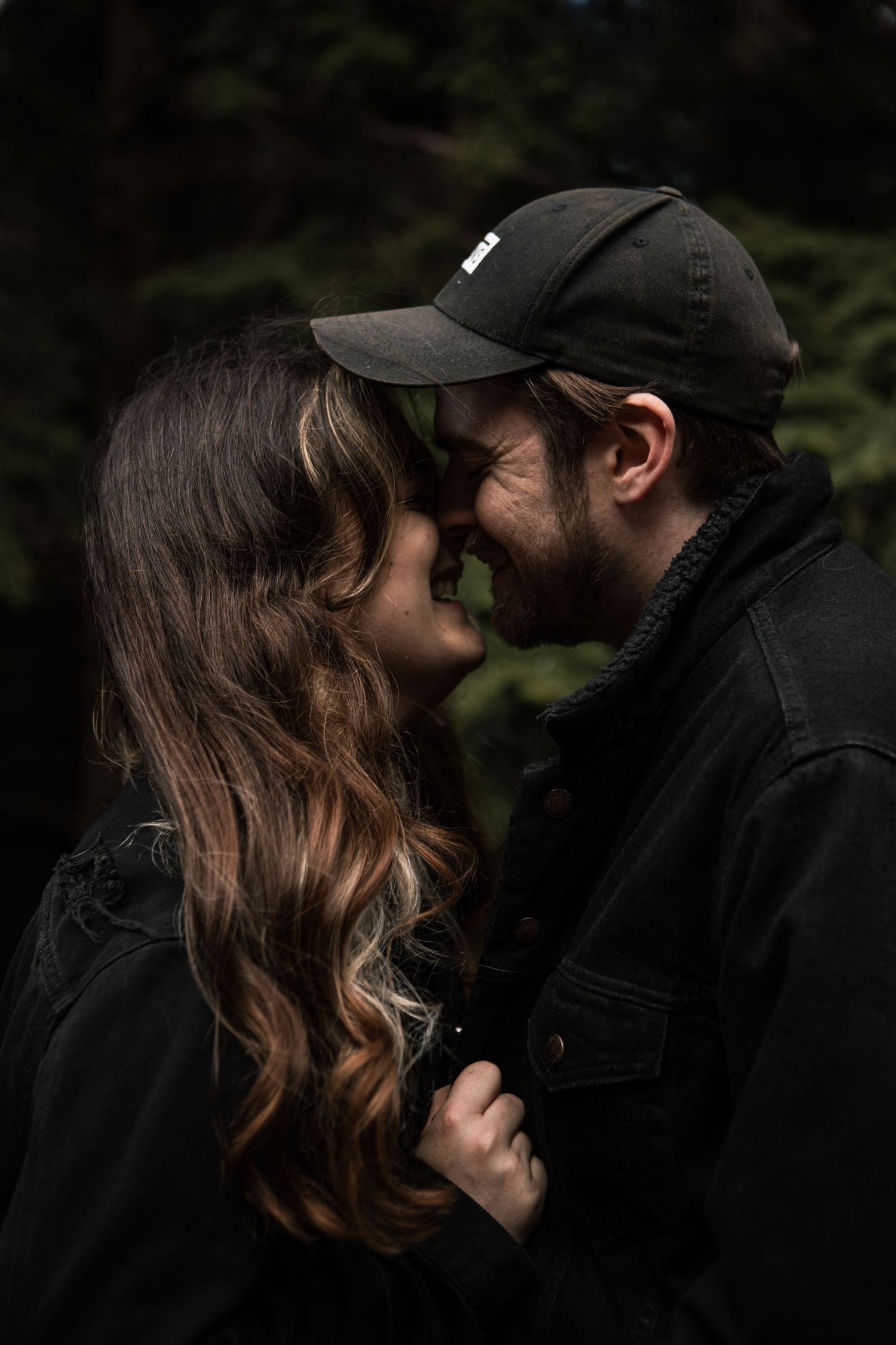 Couple sharing an intimate moment outdoors, man in black cap and jacket with woman with long wavy hair.