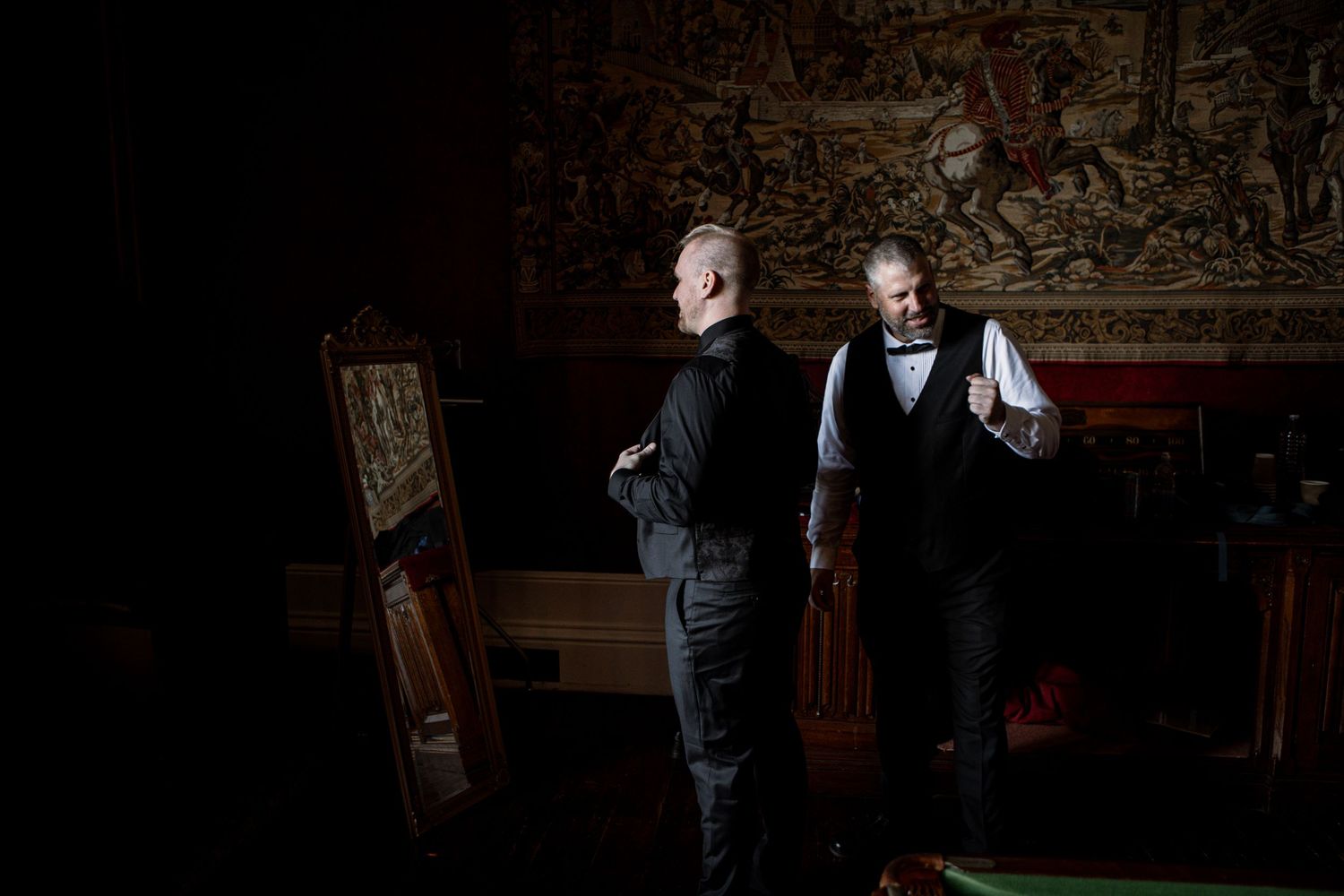 A person in a dark suit stands in an elegantly decorated room with ornate wallpaper and vintage furnishings.