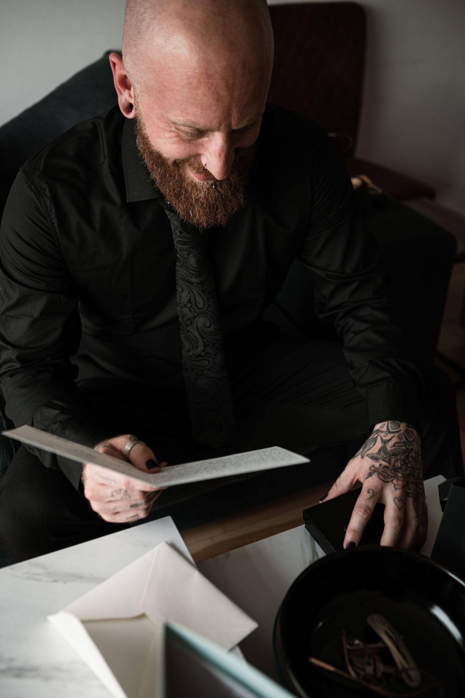 Bald bearded tattooed man in black shirt reading a document at a desk with papers and a dark bowl.