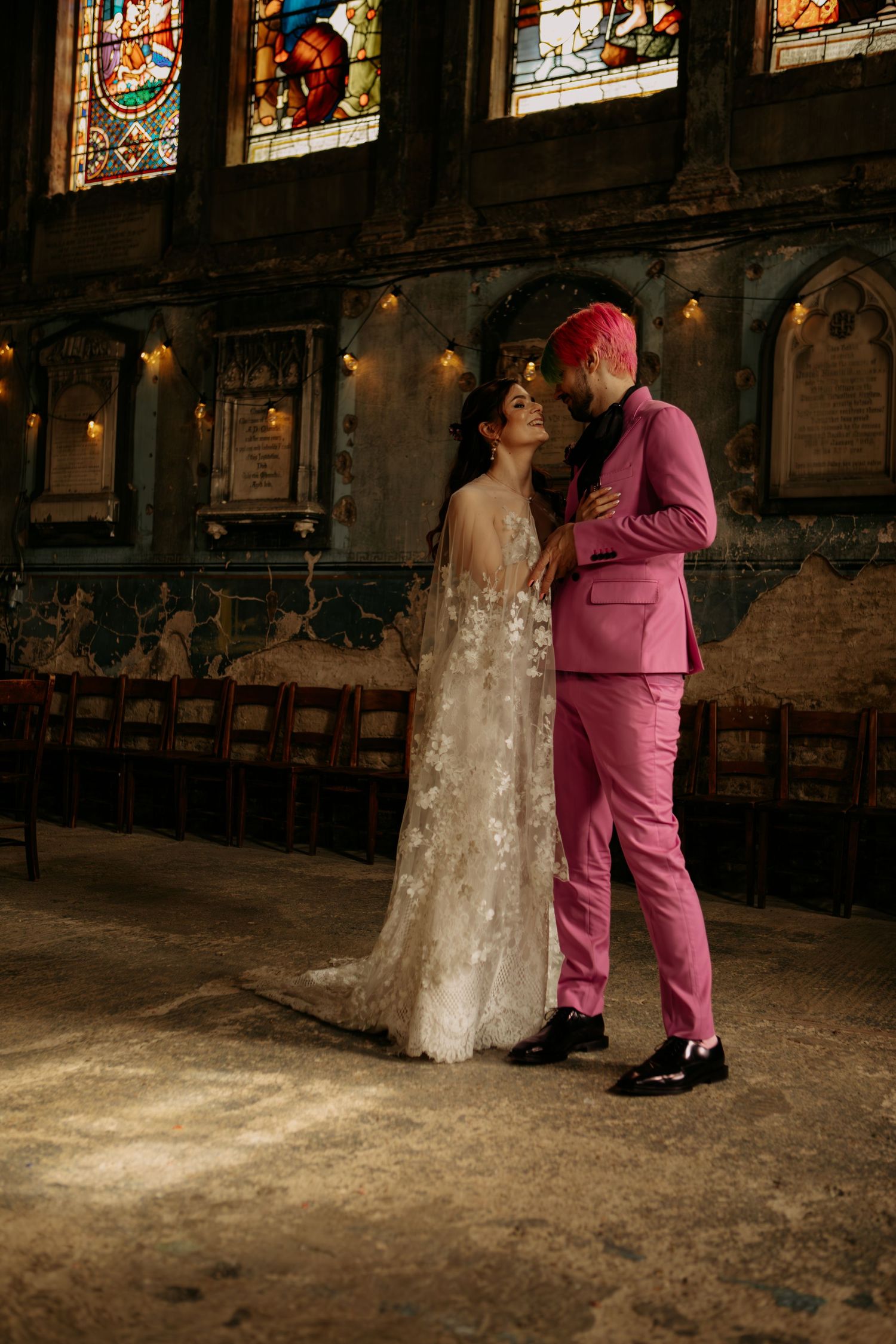 Bride in floral gown and groom in pink suit share romantic moment in ornate church with stained glass windows.