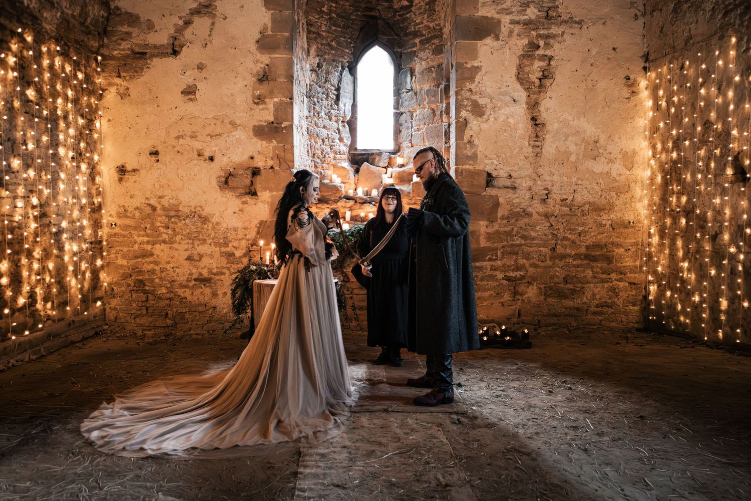 Gothic wedding ceremony in a candlelit stone ruin, bride in flowing gown with officiant and groom in dark medieval attire.