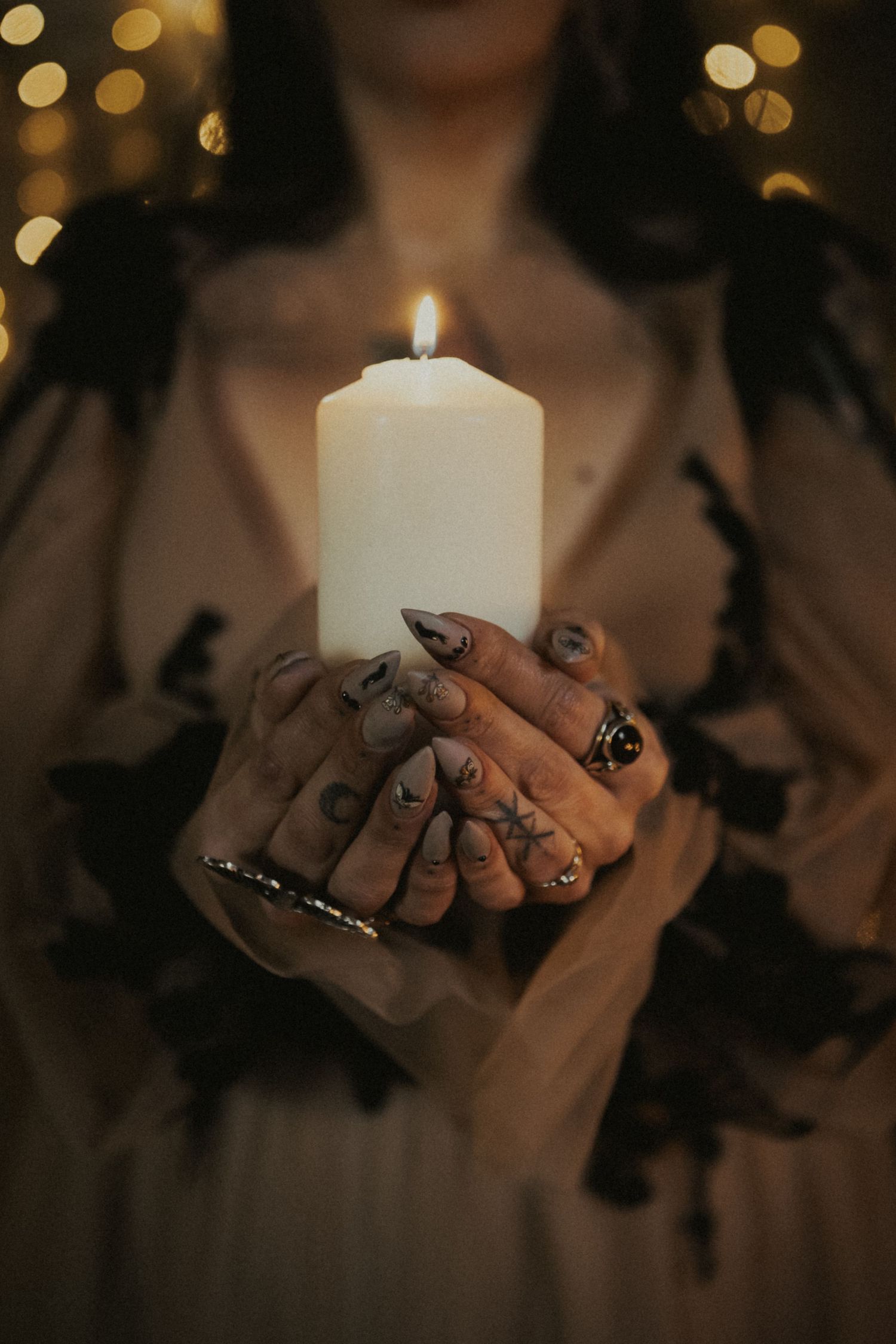 Hands with dark nail art and rings holding a lit white pillar candle against a bokeh light background.
