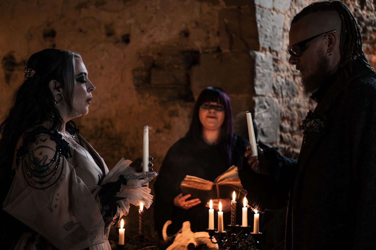 Gothic bride and groom exchange vows by candlelight with officiant reading from book in dark stone ceremony space.