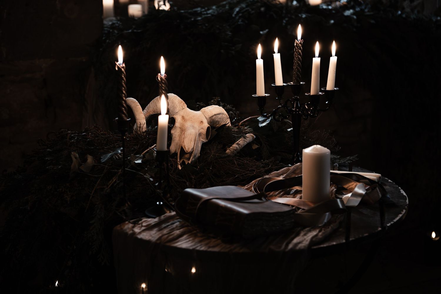 Dark gothic altar with ram skull, black candelabra, lit candles, and a black book on a wooden table.