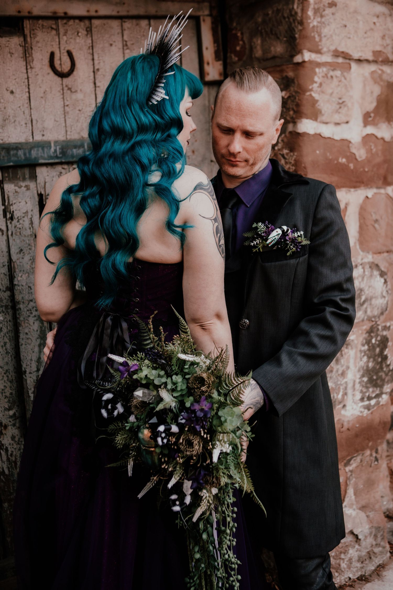 Gothic bride with teal hair and purple gown holds wild bouquet beside groom in black suit outdoors.