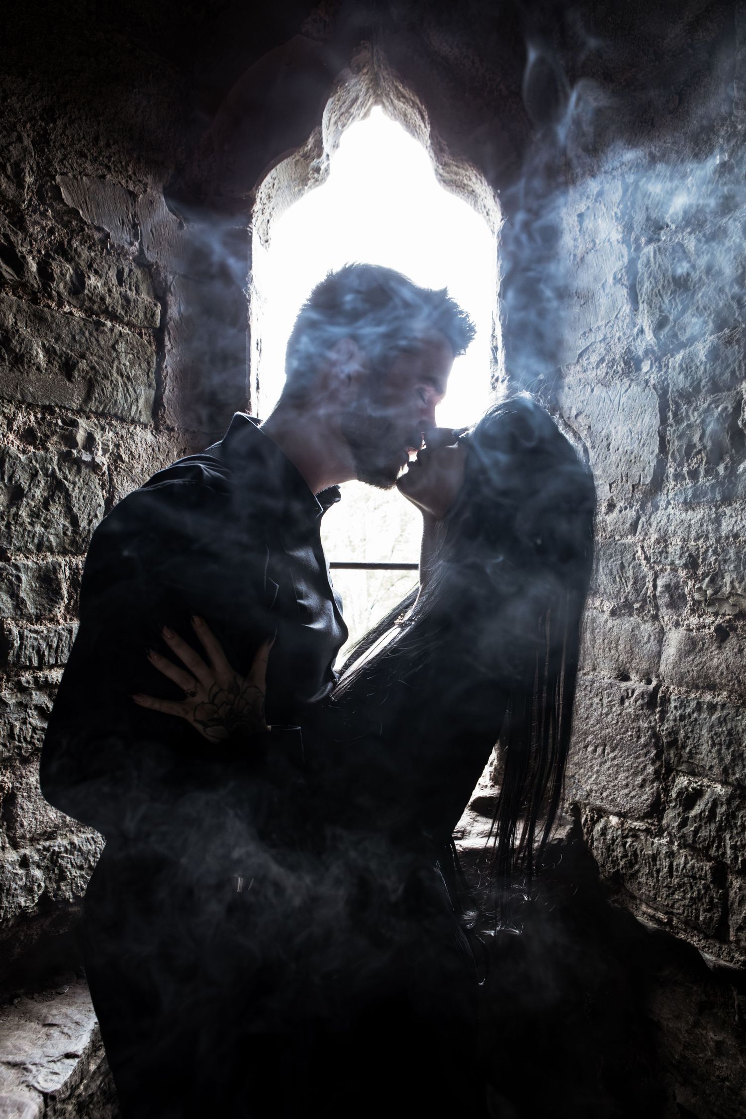 Romantic couple silhouetted against gothic arch window in misty stone castle, sharing intimate moment.