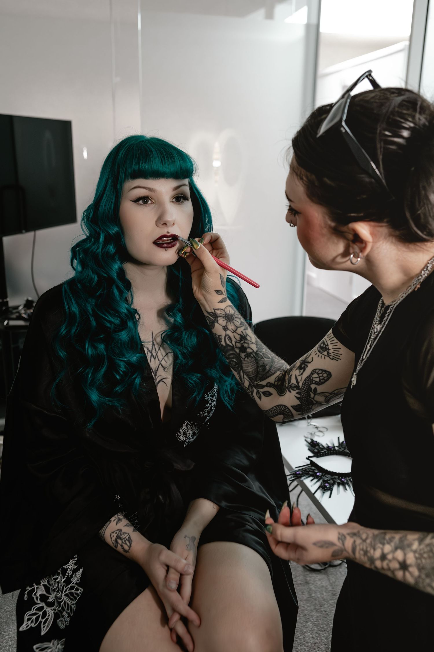 A makeup artist with tattoos applies lipstick to a woman with teal curly hair wearing a black satin robe.