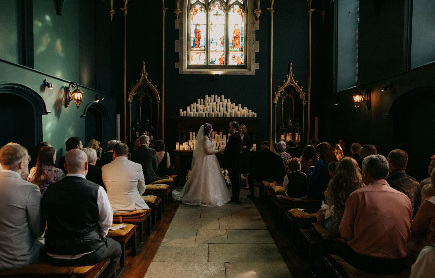 Elegant church wedding ceremony with bride and groom at altar, candlelit stained glass backdrop, guests seated in wooden pews.