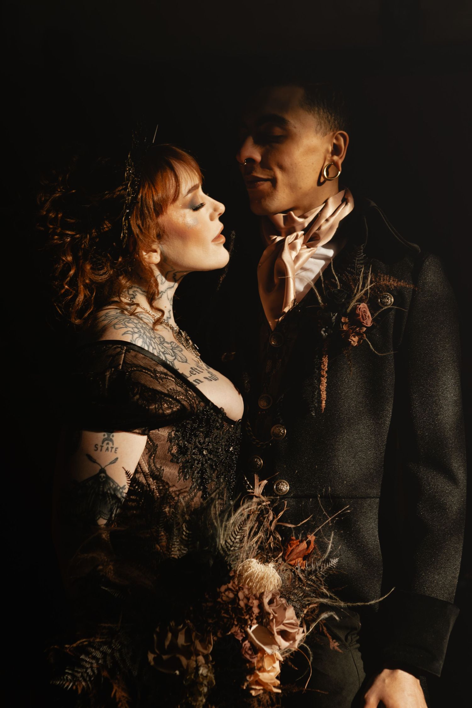 A couple in gothic wedding attire poses dramatically against a dark background, the bride holding dried botanical bouquet.