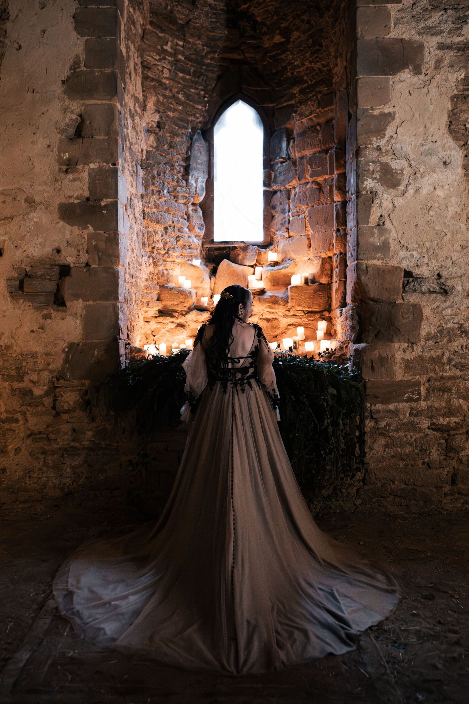 Bride in elegant dark gown standing before candlelit gothic stone arch window in ancient castle ruins.