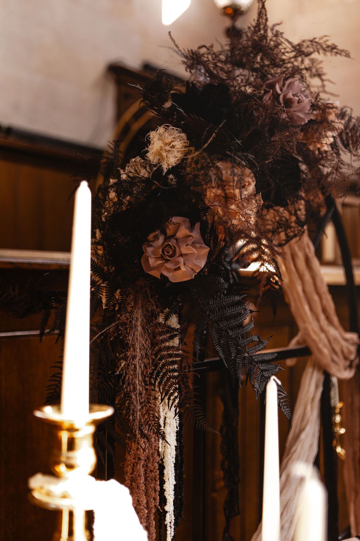 Dark gothic wedding floral arrangement with dried black ferns, mauve blooms, and candlestick in moody church setting.