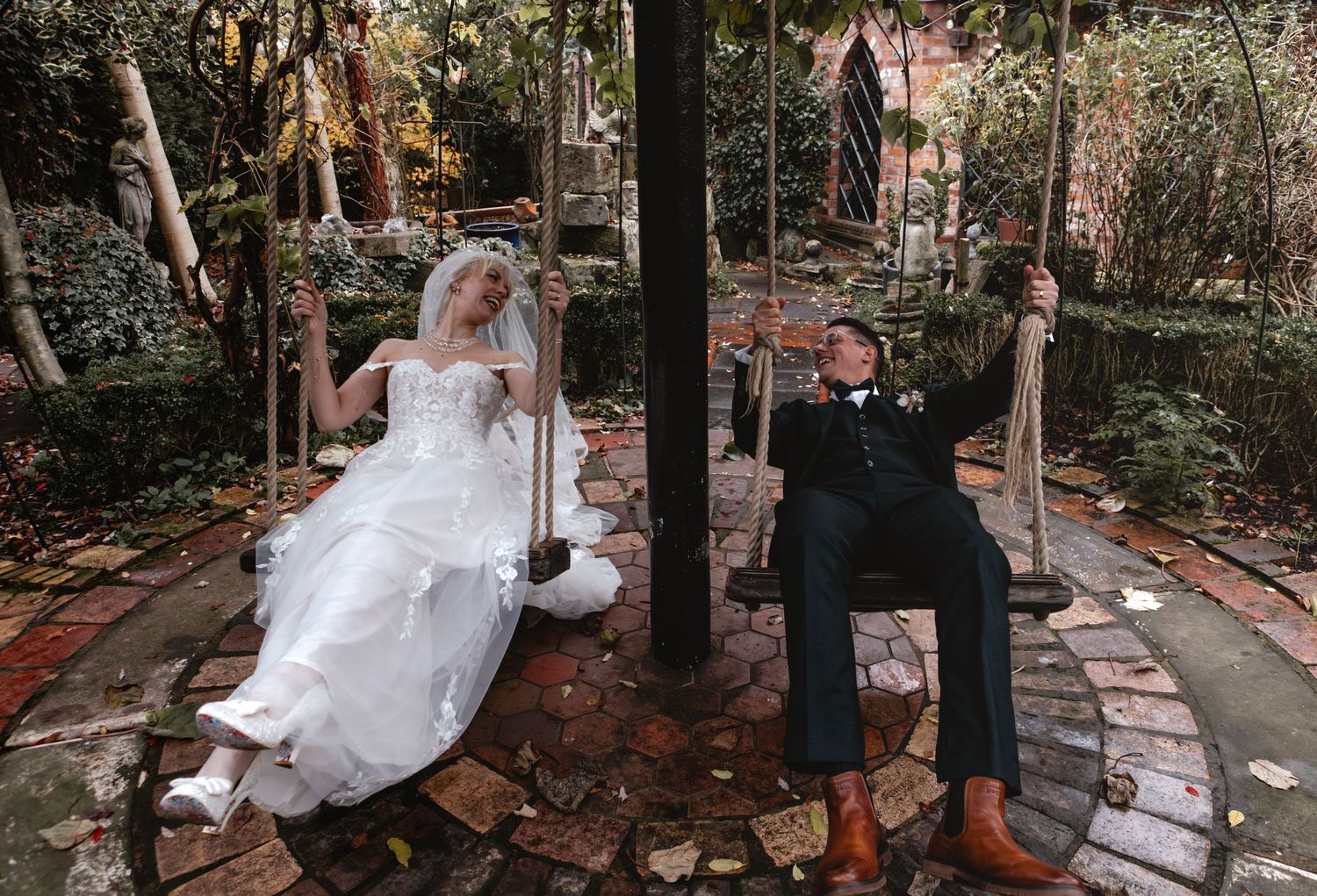 Bride and groom sitting on garden swings in lush outdoor setting, wearing wedding attire, sharing a romantic moment.