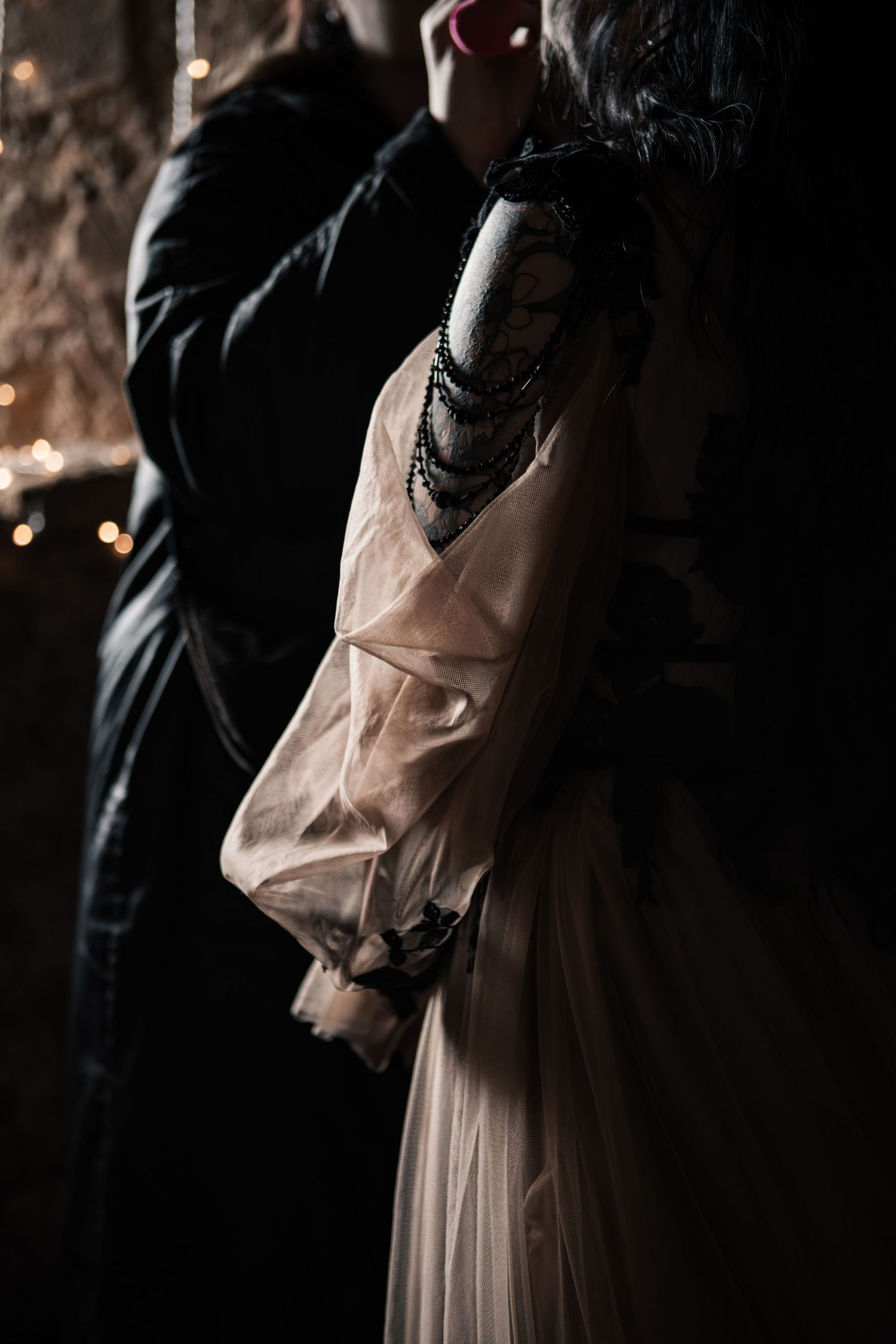 Close-up of gothic bride in sheer champagne gown with black bead accessories held by groom in dark stone venue.