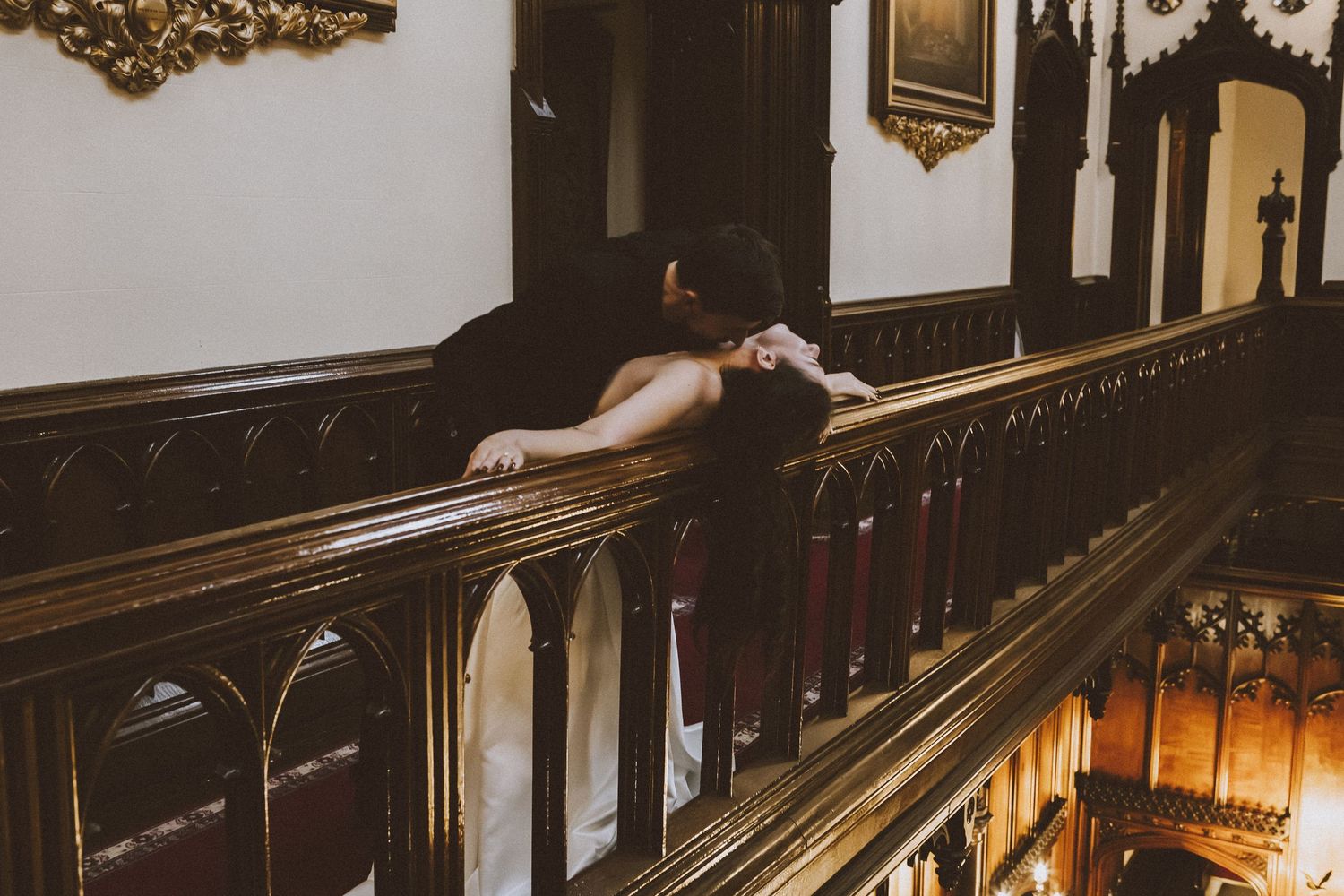 Couple sharing a romantic kiss on an ornate wooden balcony railing inside a historic building.