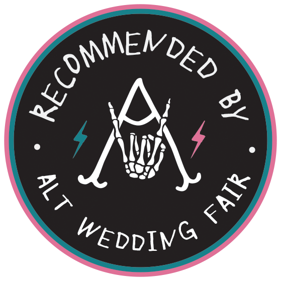 A circular black logo with pink and blue borders featuring a skeleton hand making a peace sign between crossed canes.