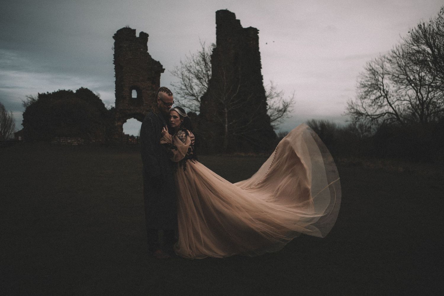 Couple embracing at dusk near ancient castle ruins, bride's flowing champagne gown billowing dramatically in the wind.