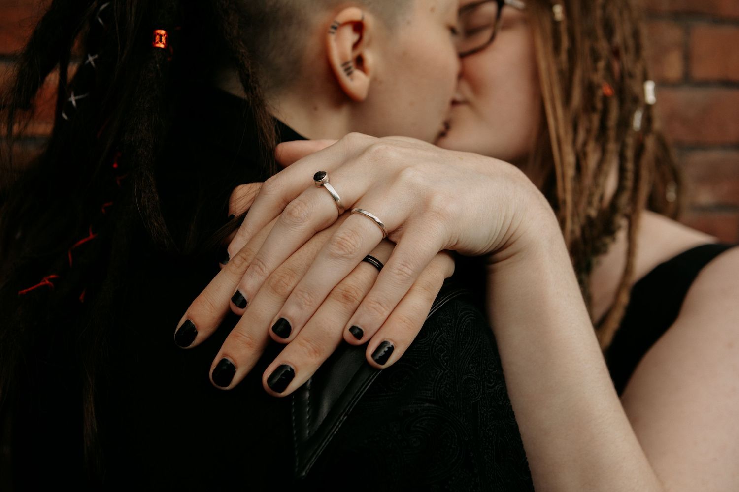 Two people sharing a kiss while a hand with black nails and silver rings is displayed in the foreground.