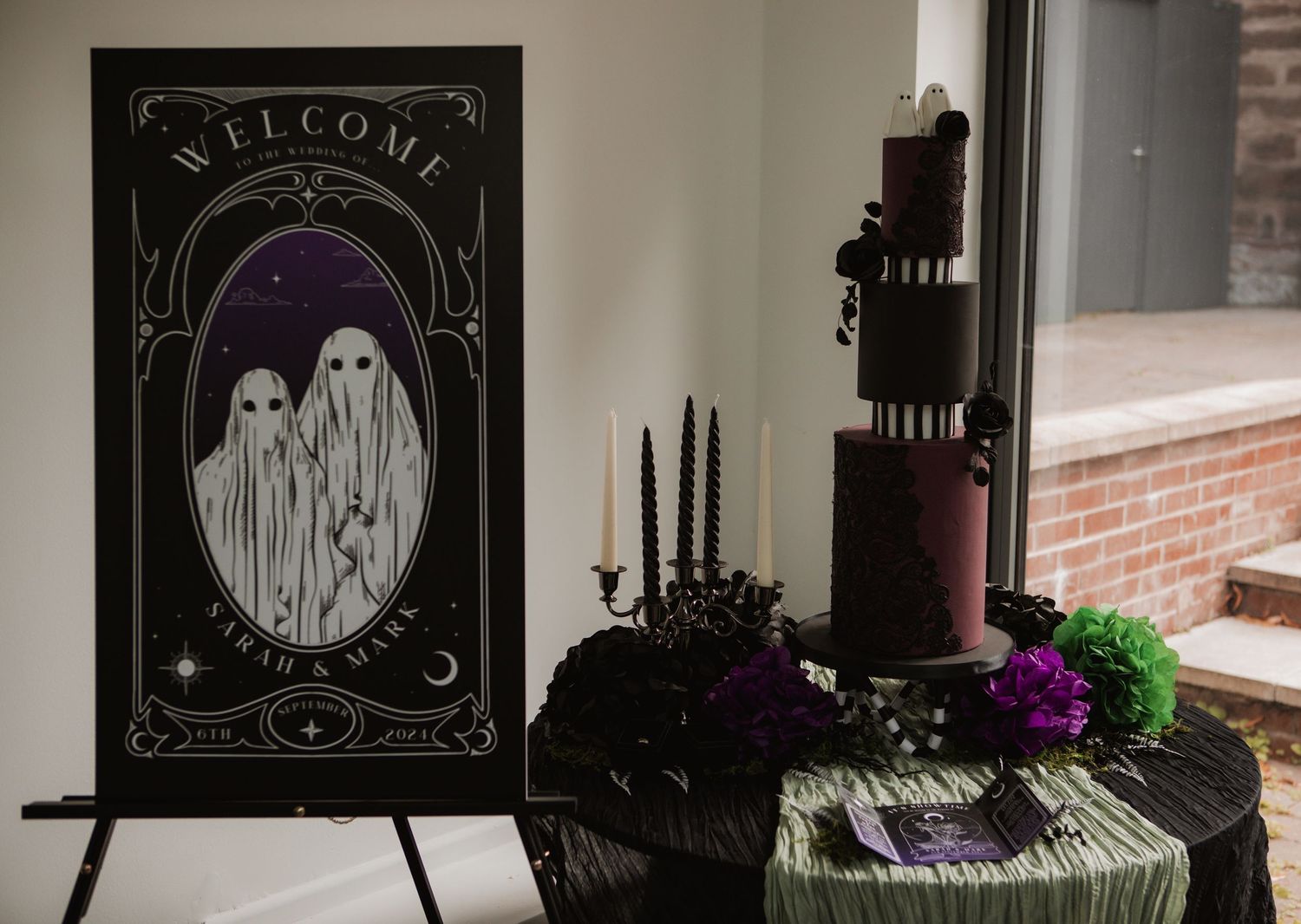 Gothic wedding display with ghost welcome sign, black candles, dark purple tiered cake, and colorful floral decorations.