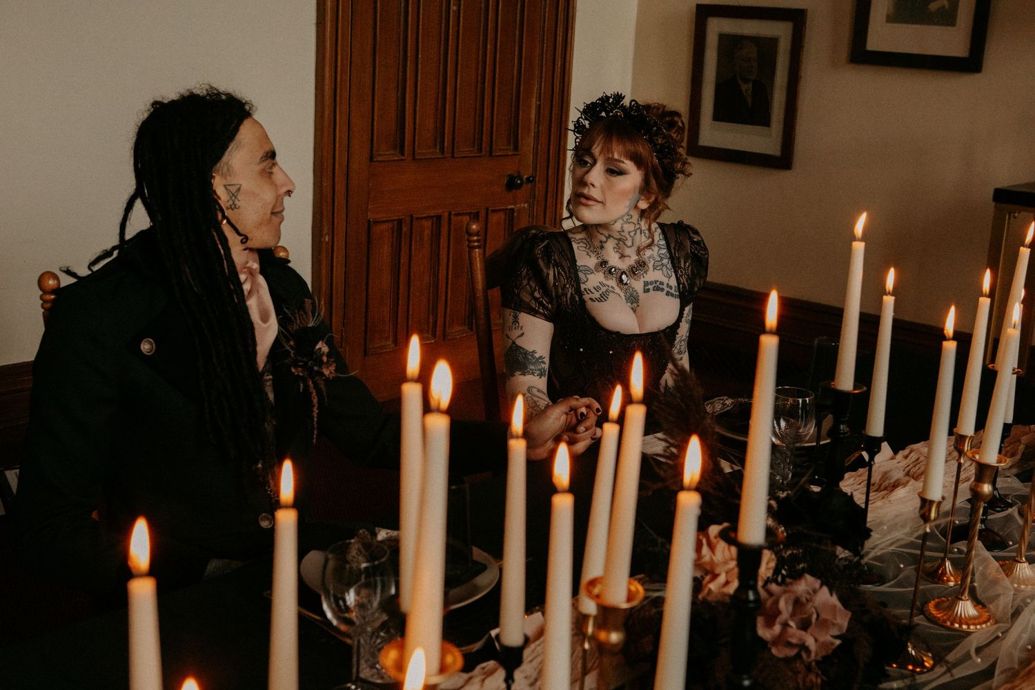 Two women with tattoos and gothic style stand by candlelit wedding reception table with tall white candles.