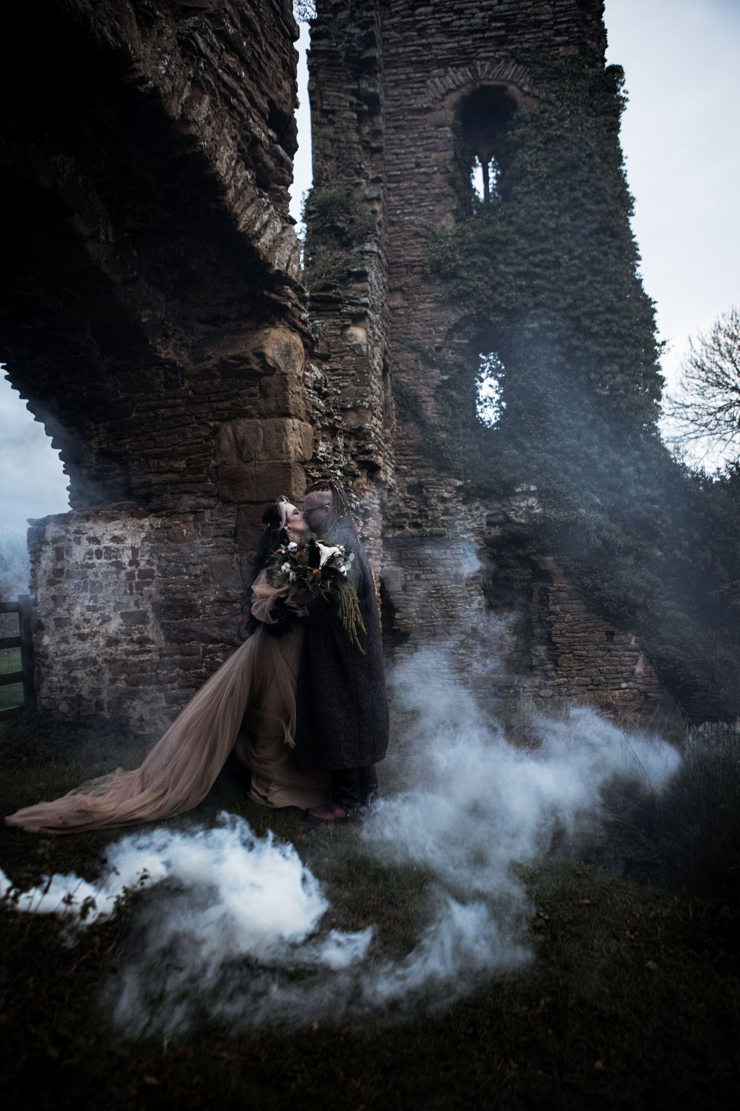 Gothic wedding couple embracing at ancient stone castle ruins surrounded by dramatic white smoke.