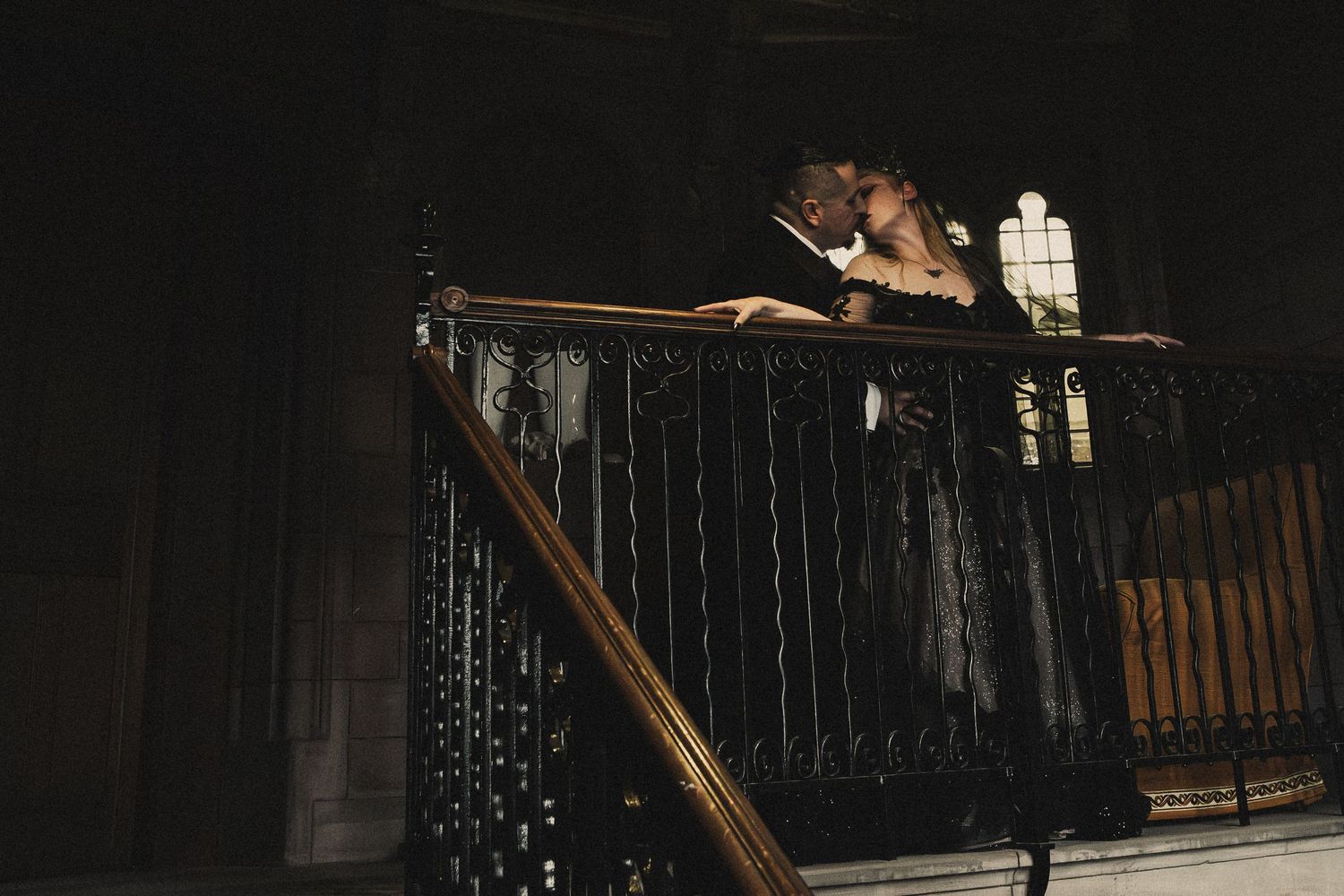 Wedding couple sharing a kiss on ornate iron staircase balcony in dimly lit historic venue.