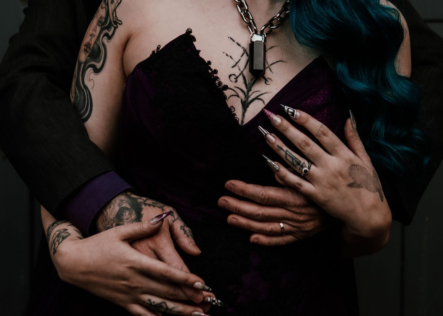 Couple embracing, woman with blue hair, tattoos, purple corset, long nails, and silver necklace in gothic style.