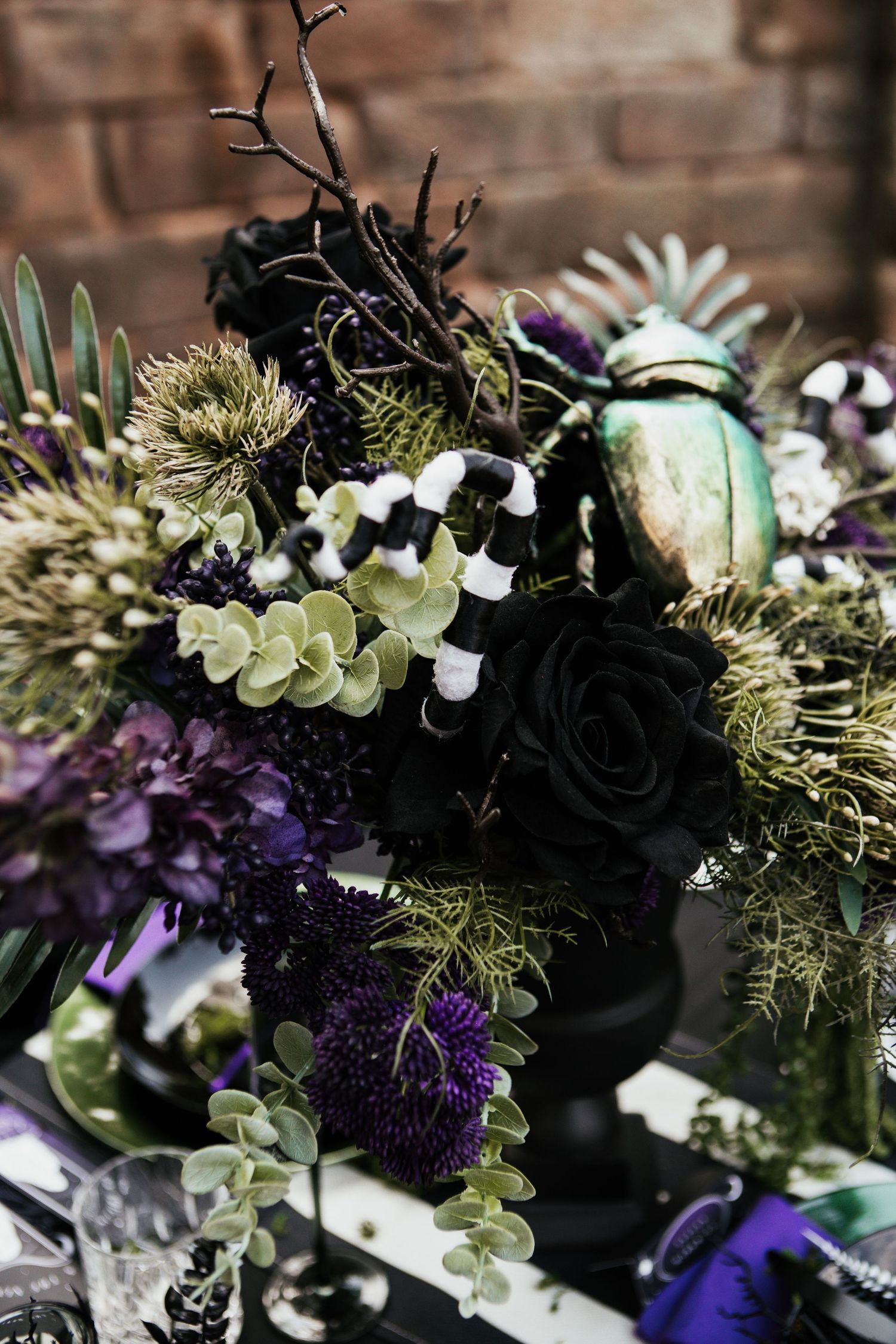 Dark gothic floral arrangement with black roses, purple blooms, green foliage, and decorative branches in a black vase.