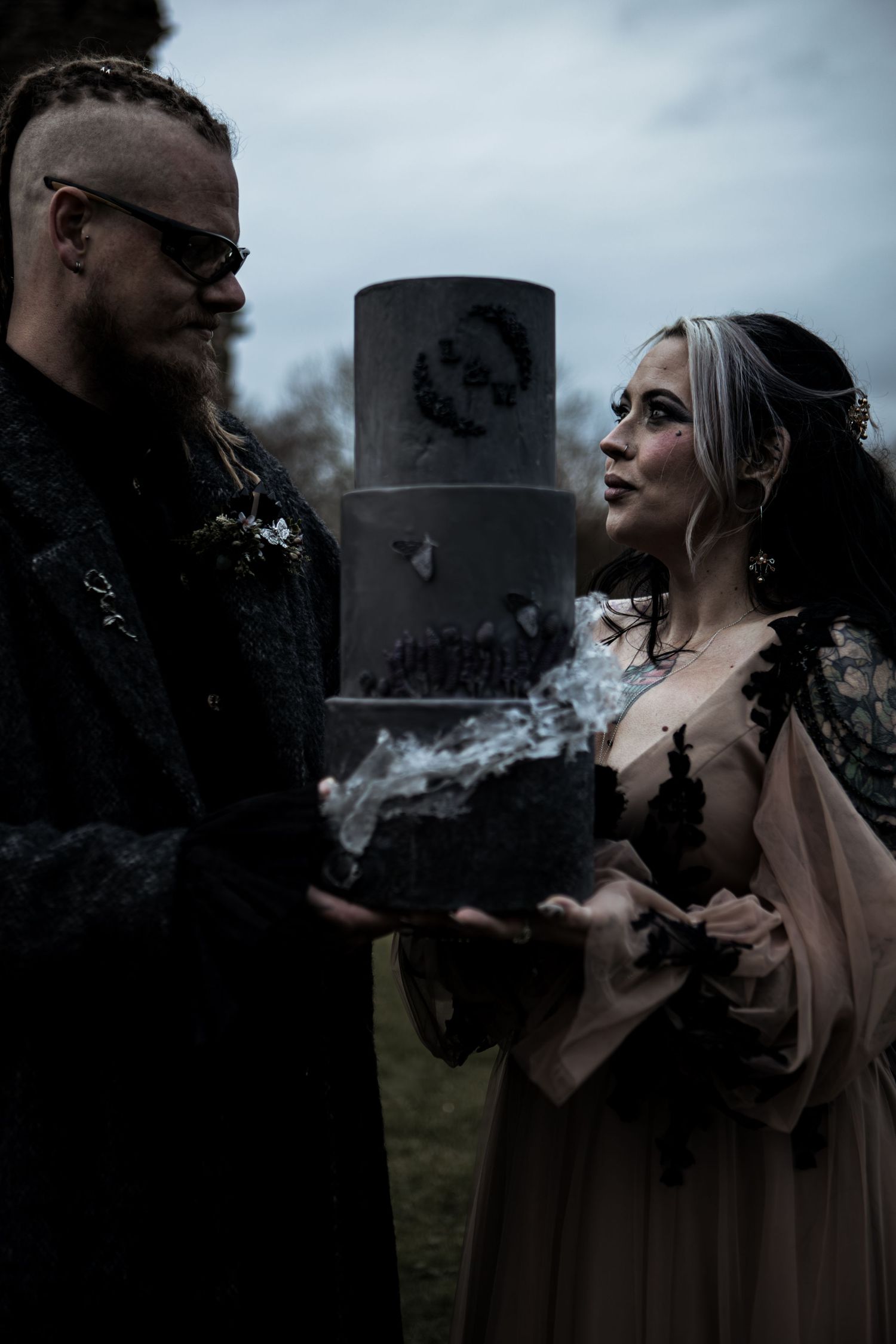 Gothic wedding couple in dark attire holding a rock, with a black tiered cake in the background.