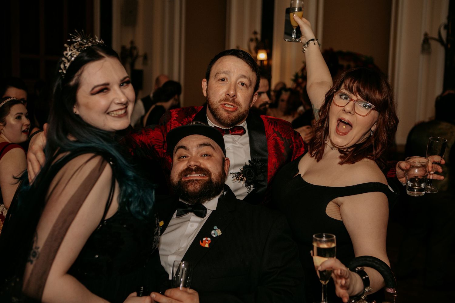 Formal party guests in elegant attire pose together while celebrating and holding drinks at an indoor event.