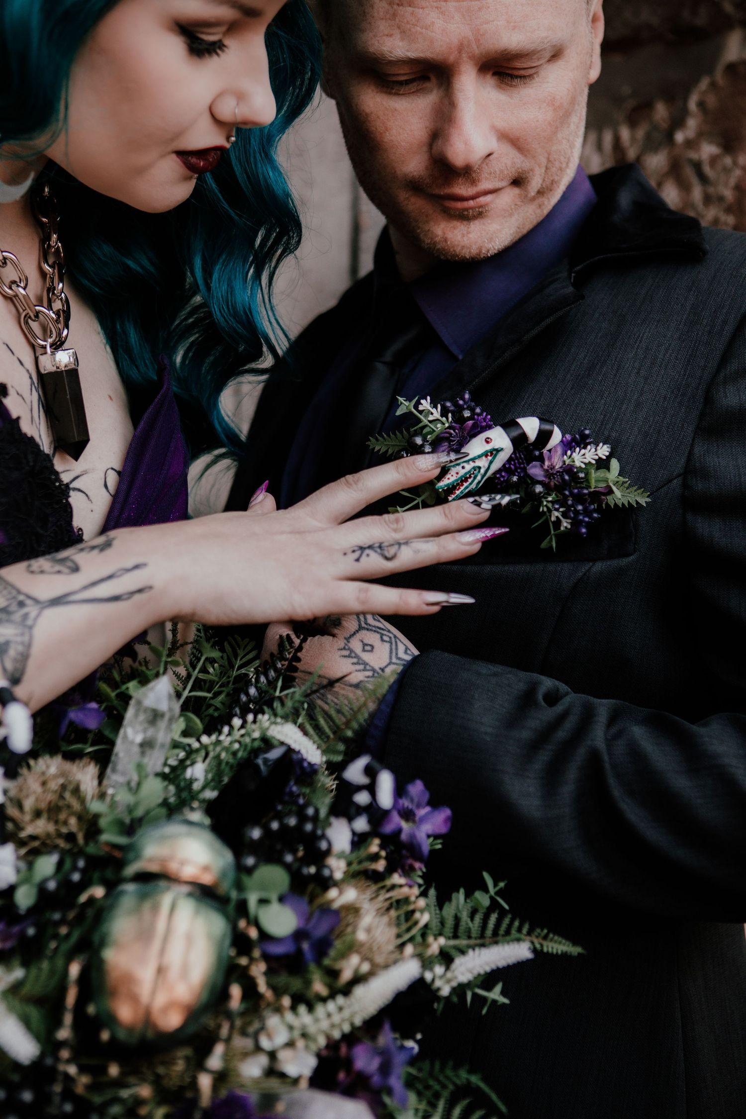 Gothic bride with teal hair and tattoos adjusting groom's boutonniere, holding dark floral bouquet with crystals.