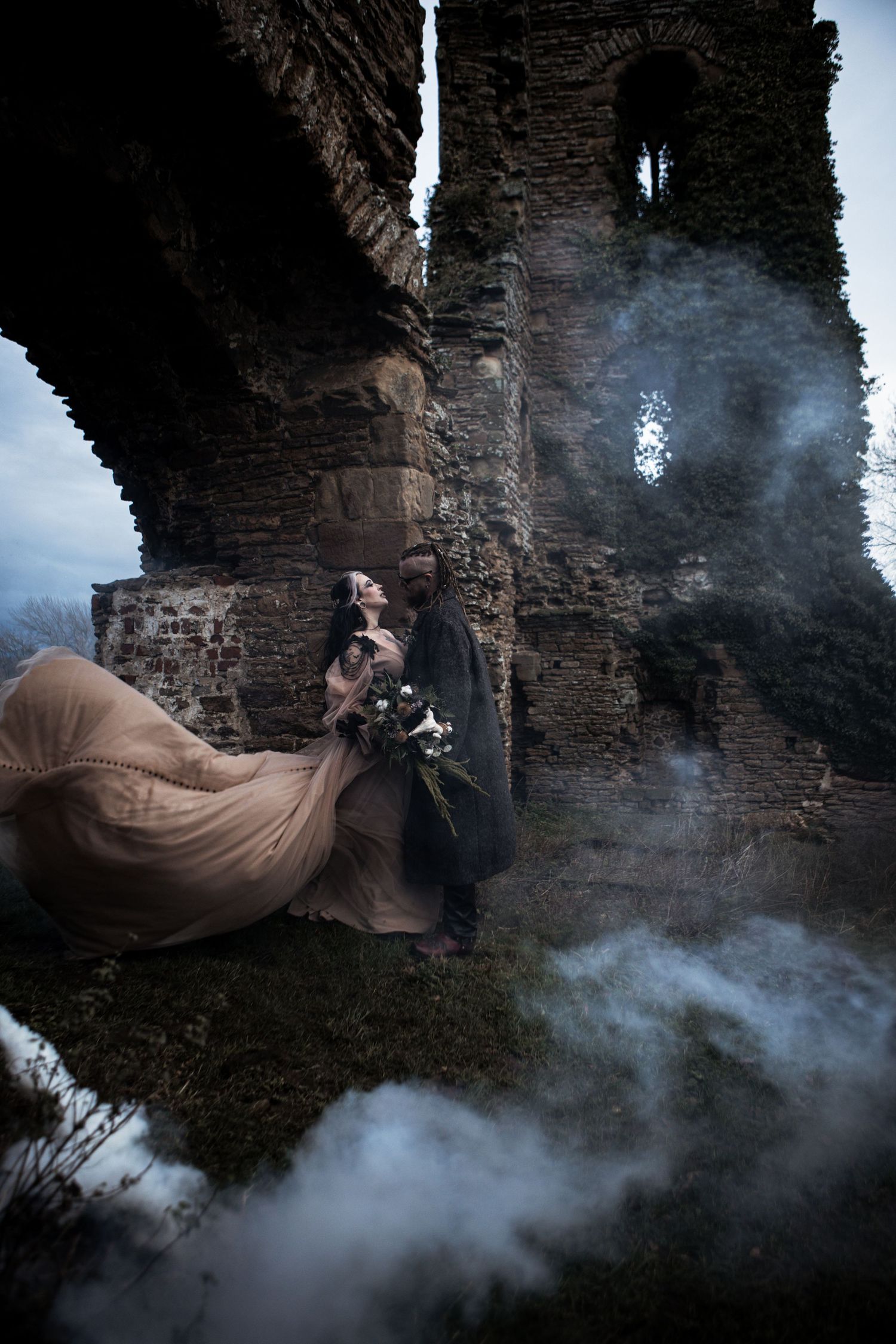 Gothic wedding couple in smoky ruins, bride in flowing tan gown with dramatic billowing skirt.