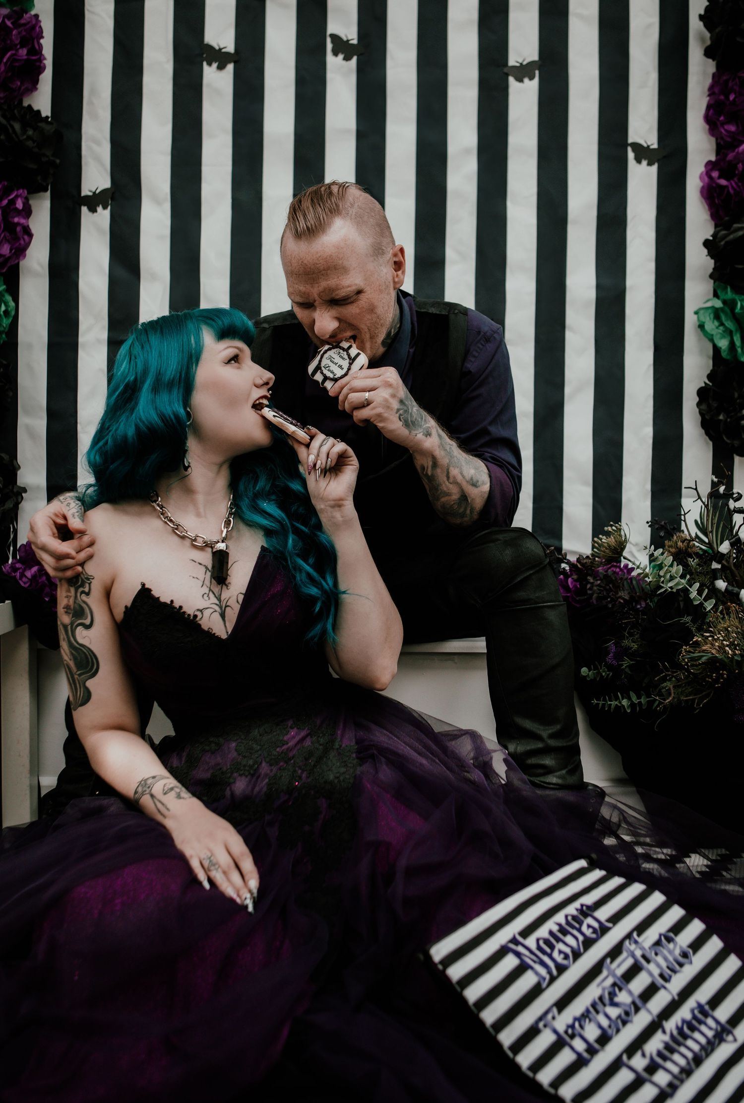 Gothic couple in Halloween-themed wedding attire sharing cake pops against black and white striped backdrop.