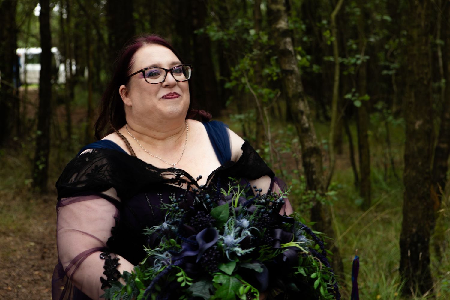 Woman in black gothic dress holding dark floral bouquet with blue thistles, standing in woodland setting.