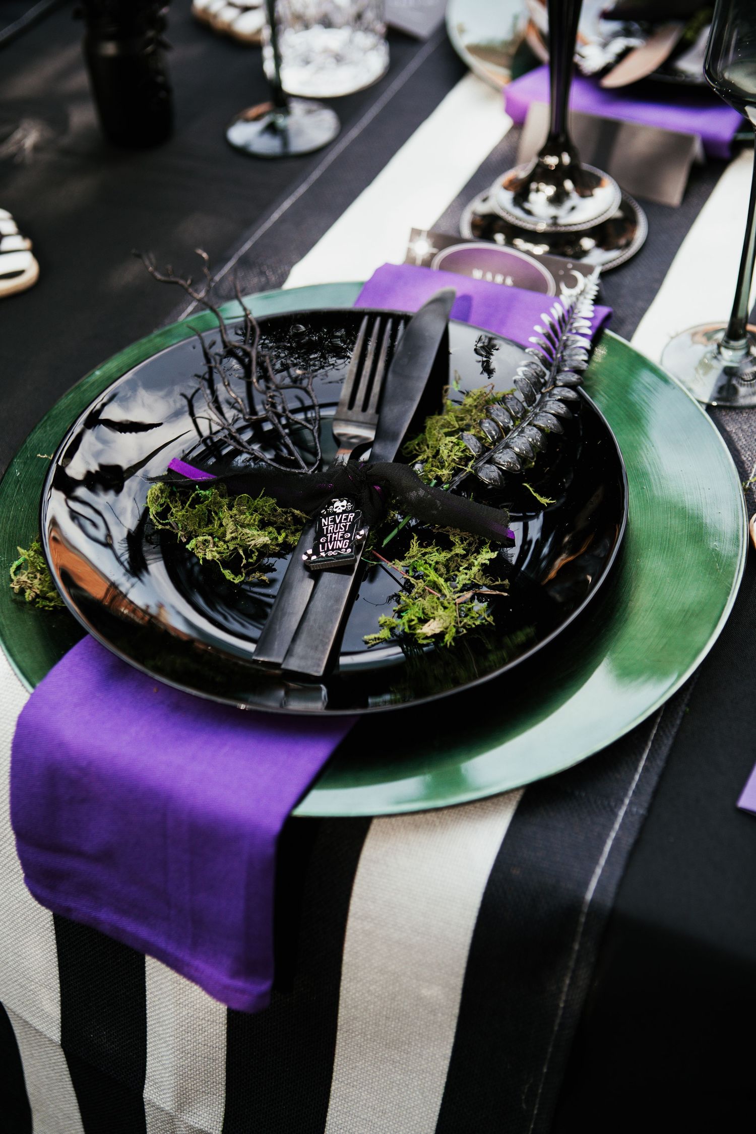Gothic Halloween table setting with black plates, purple napkins, green chargers, and dark cutlery on striped tablecloth.