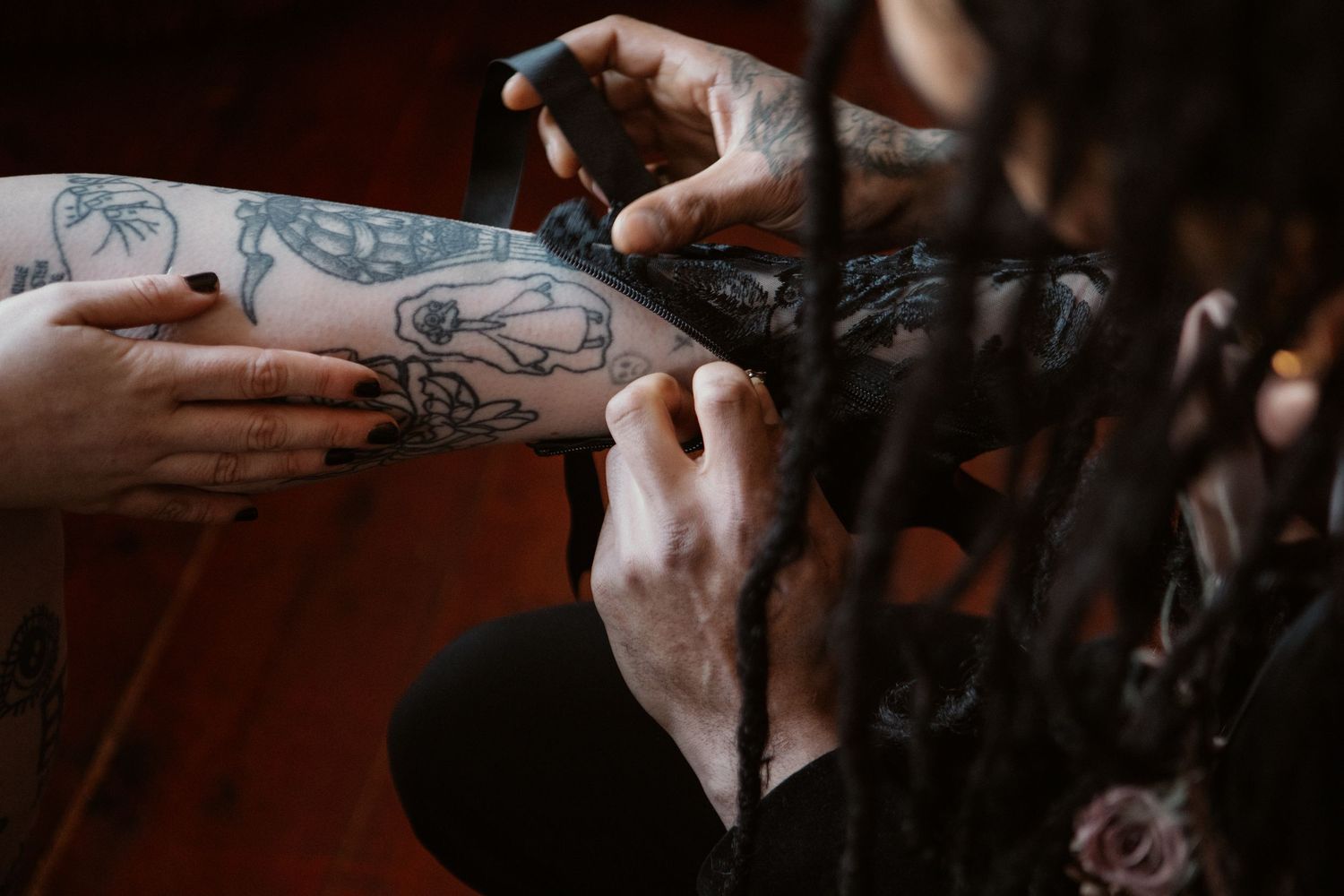 Hands with dark nails adjusting black lace lingerie over tattooed legs in dim lighting.