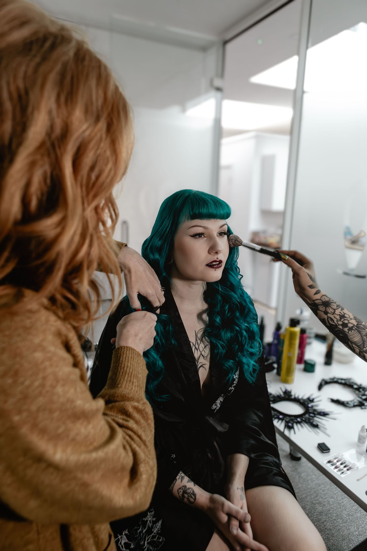 Woman with teal hair gets makeup applied by tattooed artist in a bright vanity mirror salon setting.
