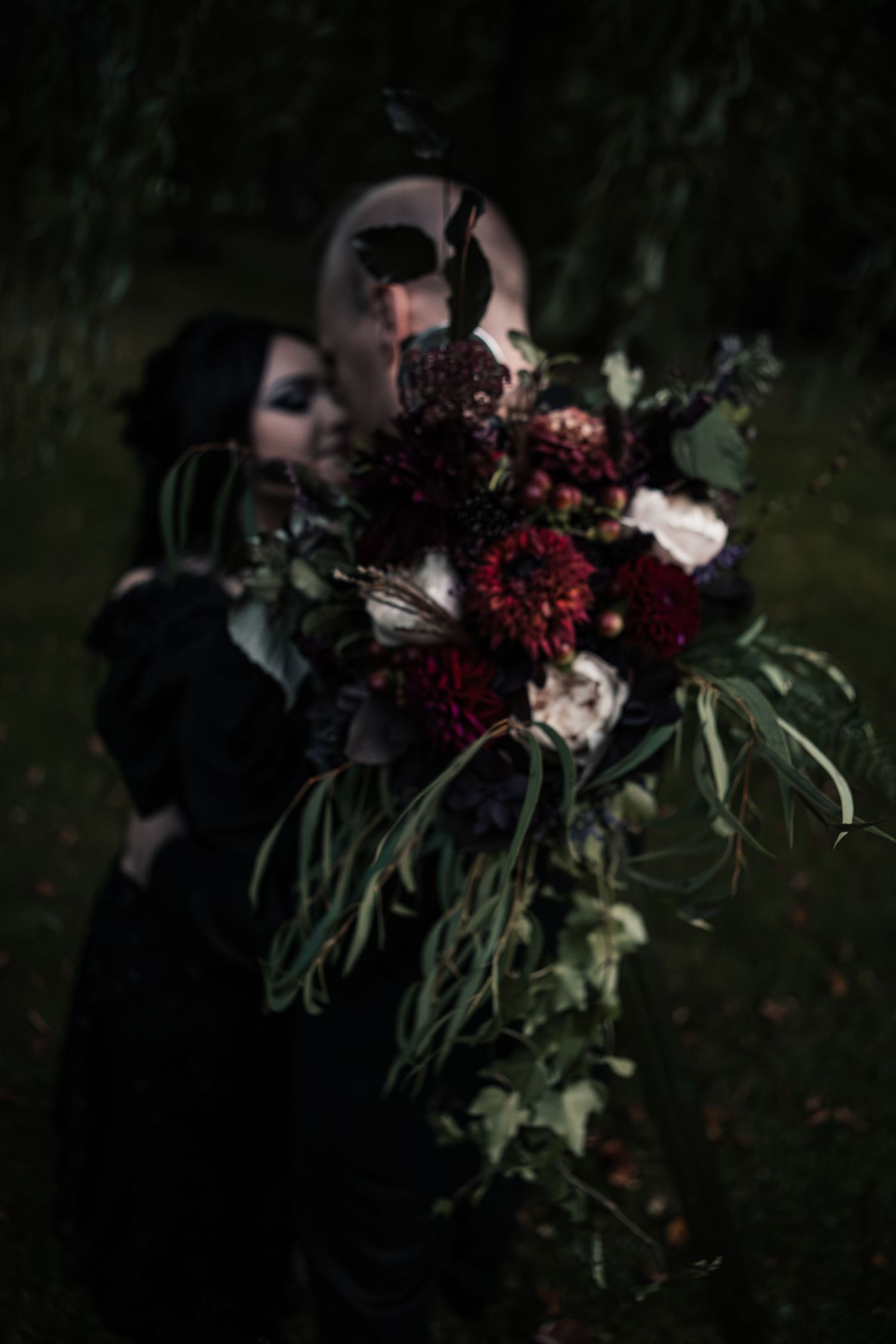 A gothic bride in black holds a lush dark floral bouquet with deep red dahlias and trailing greenery in a moody outdoor setting.
