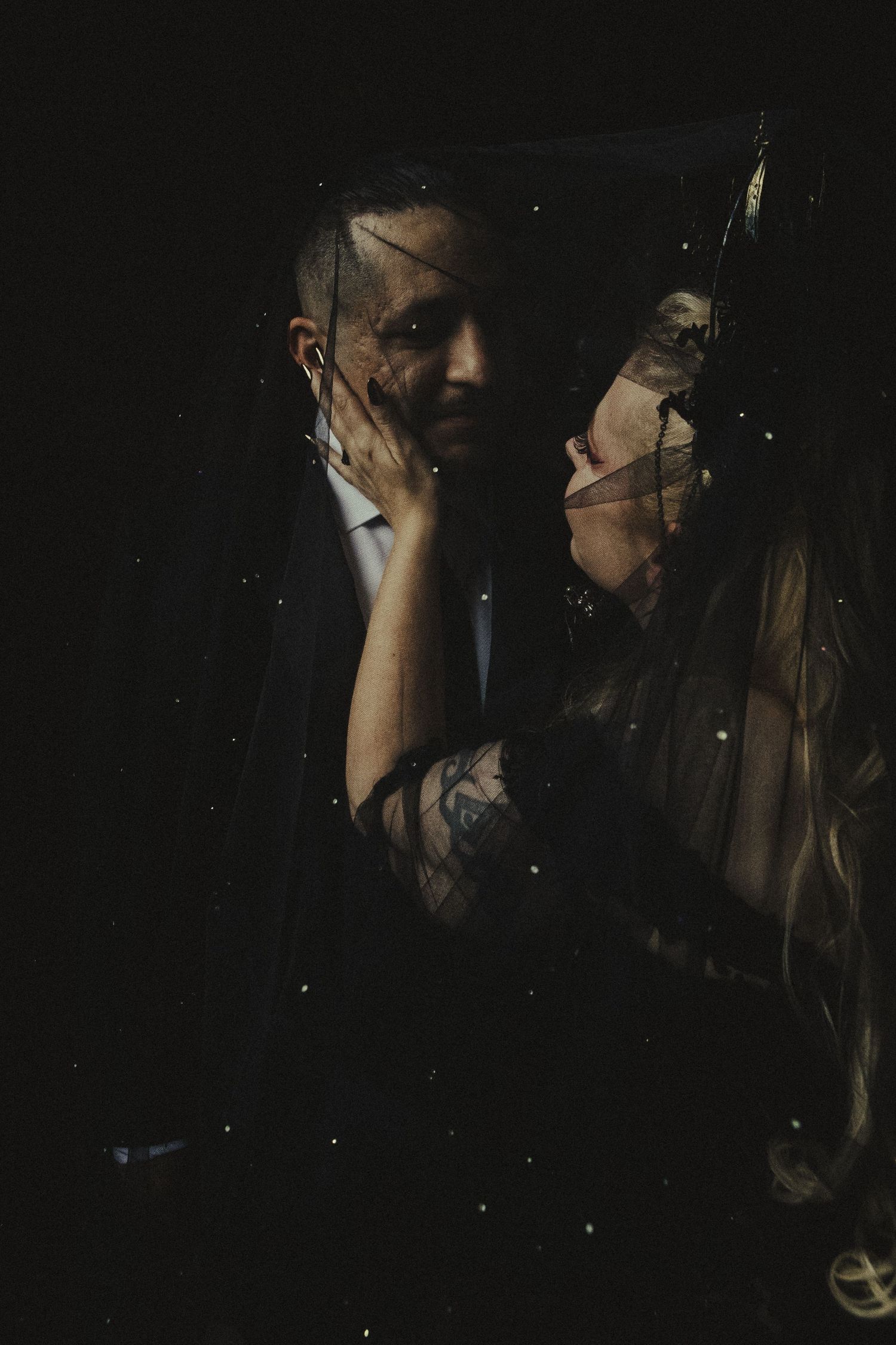 A couple shares an intimate moment under a veil in dark moody lighting, the bride's tattooed arm cradling the groom's face.
