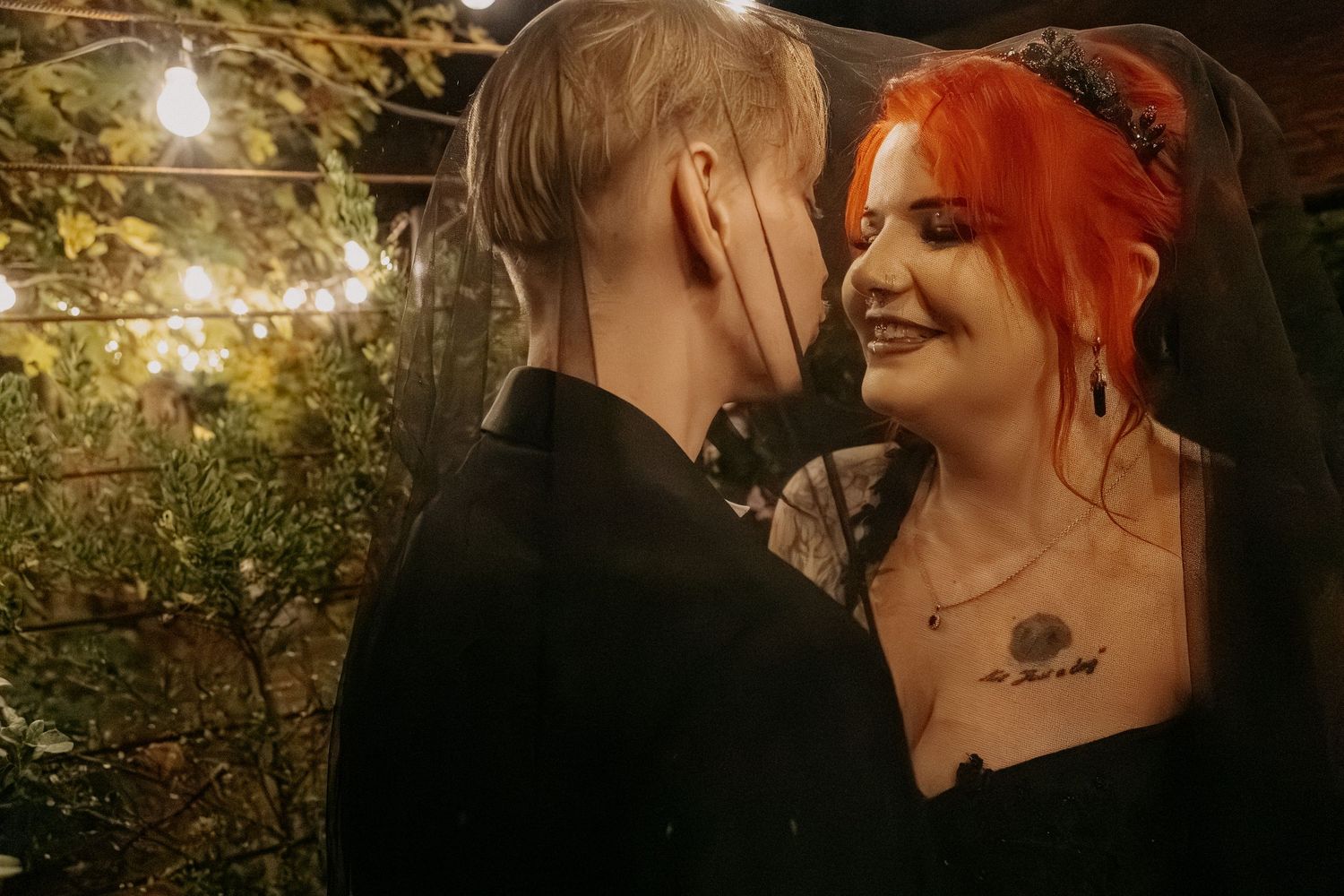 Couple in black outfits share a romantic moment outdoors at night, woman with red hair and tattoos smiling.