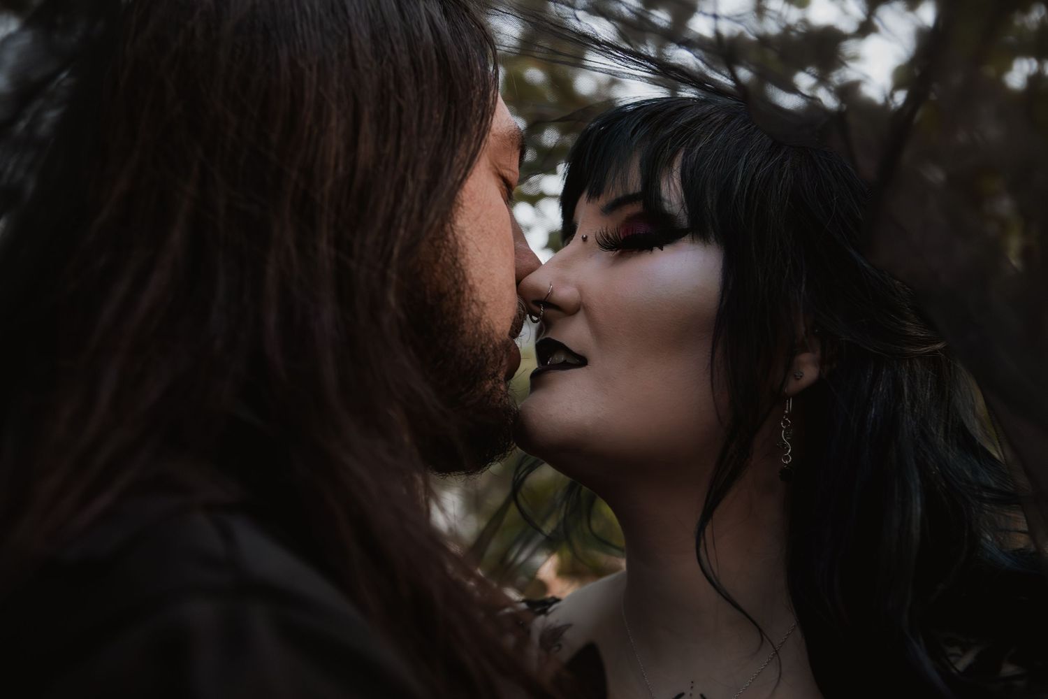 A gothic couple sharing a romantic kiss outdoors, with dark makeup and long hair against a moody, blurred natural background.