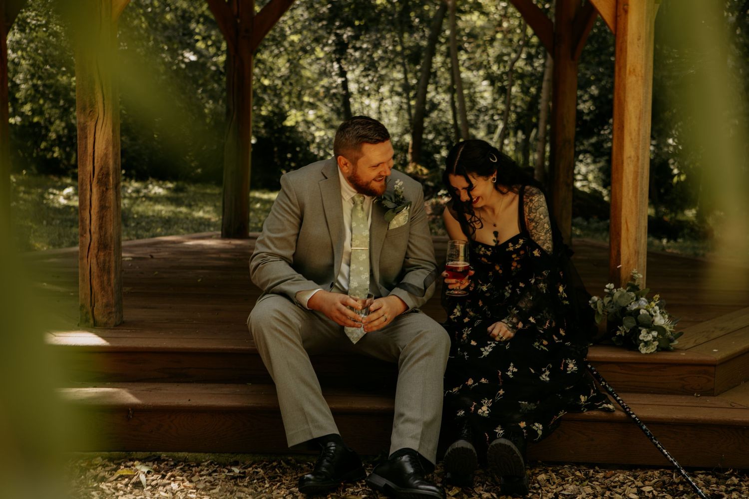 Couple in wedding attire sitting together on wooden gazebo steps, sharing a romantic moment surrounded by lush greenery.