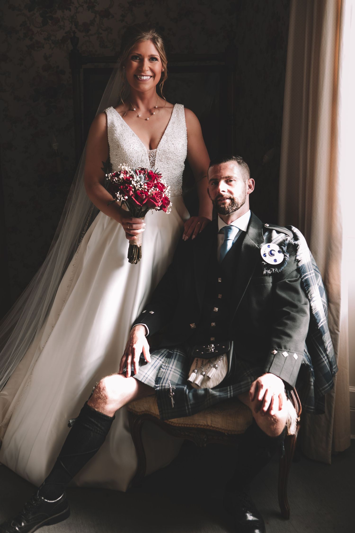 Bride in white gown holding red bouquet poses with groom in traditional Scottish kilt at wedding portrait.