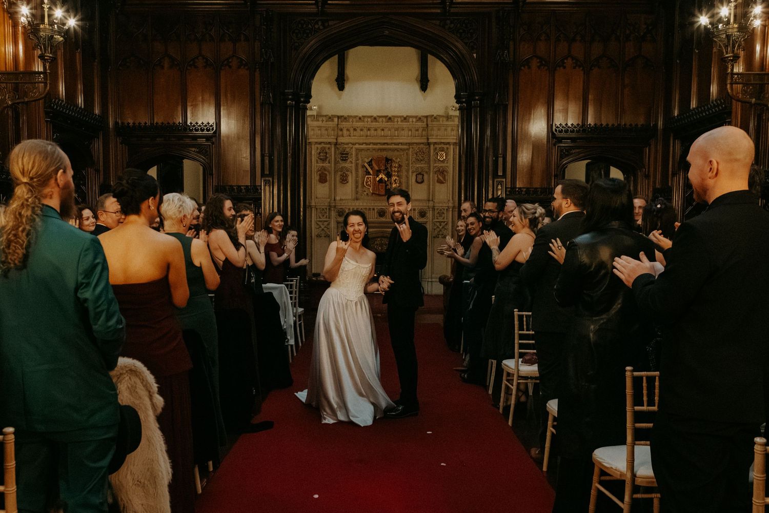 Newlyweds walk down red carpet aisle in ornate wood-paneled wedding venue as guests applaud.