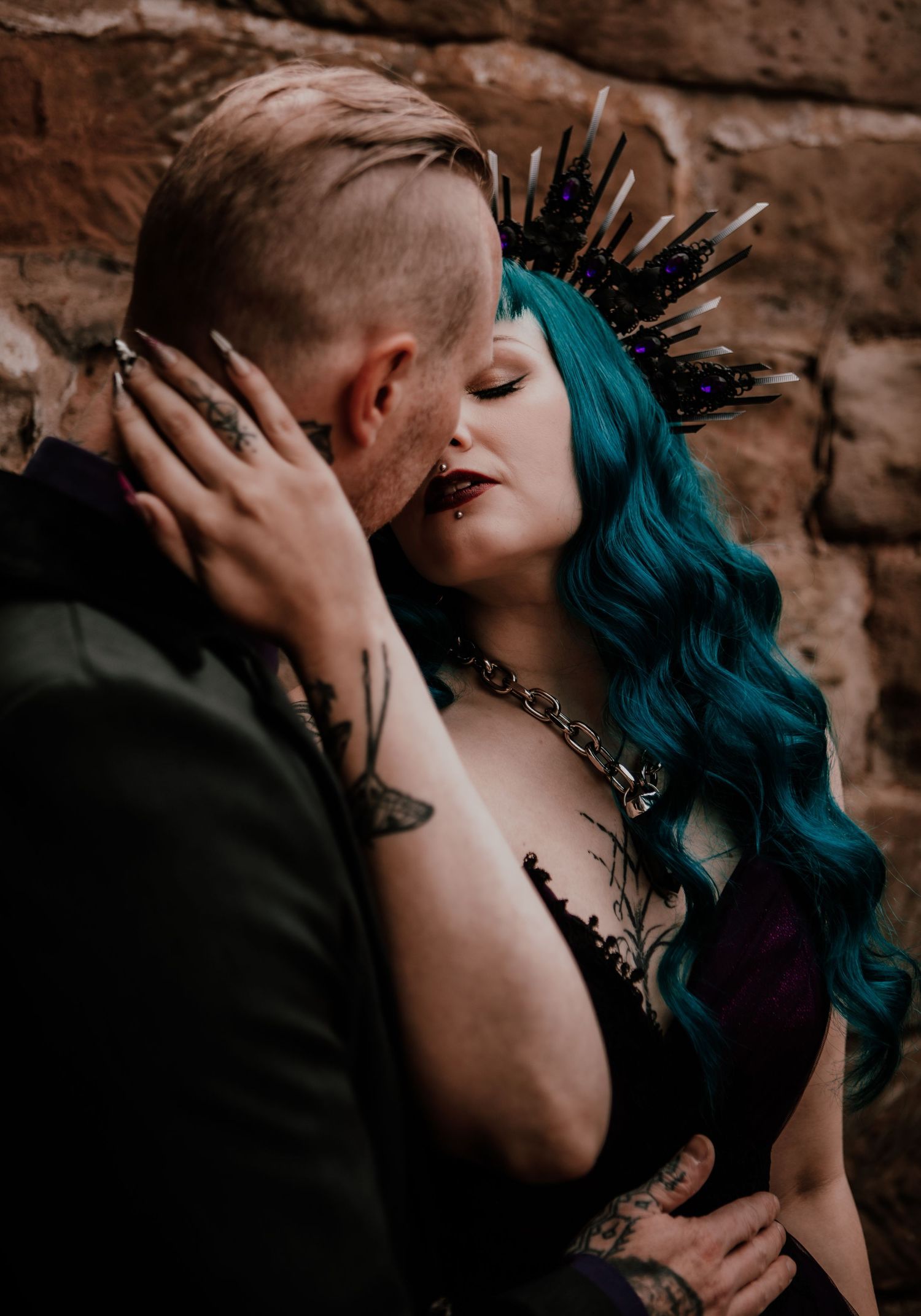 Couple sharing intimate moment, woman with teal hair and dark crown, both tattooed, against rustic stone wall.