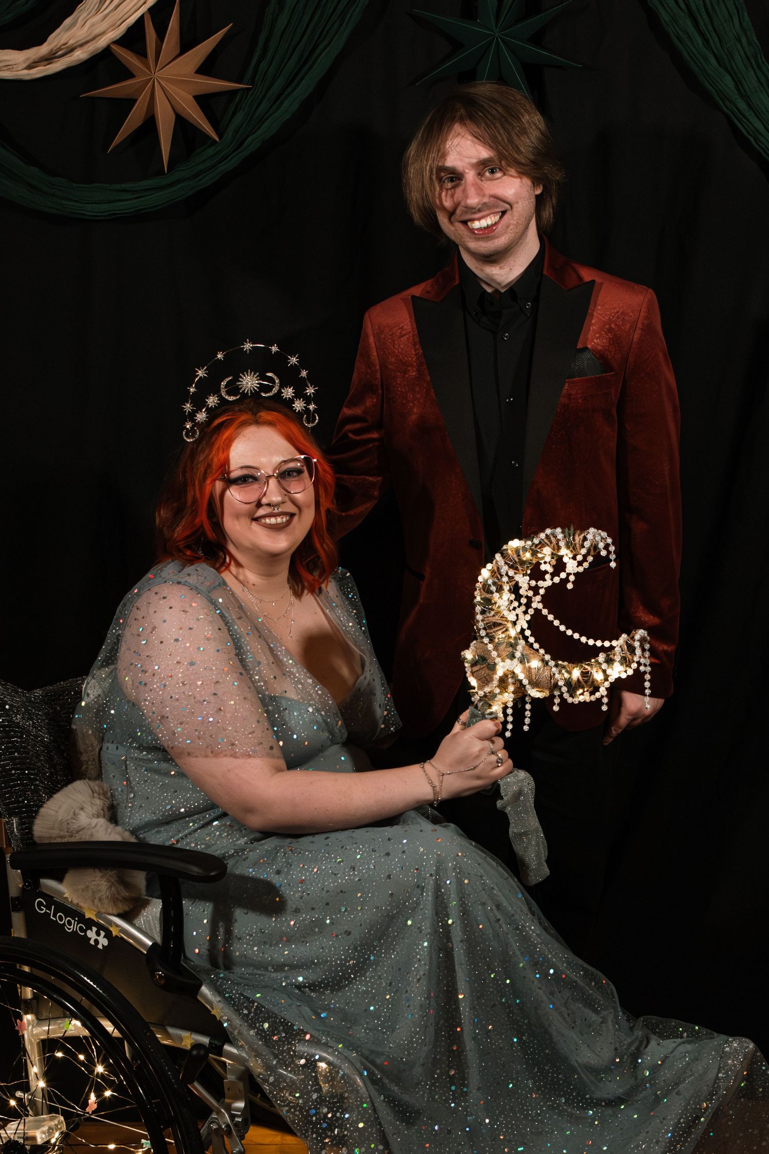 A smiling woman in a wheelchair wearing a silver gown and crown holds a light-up horse beside a man in a red velvet blazer.