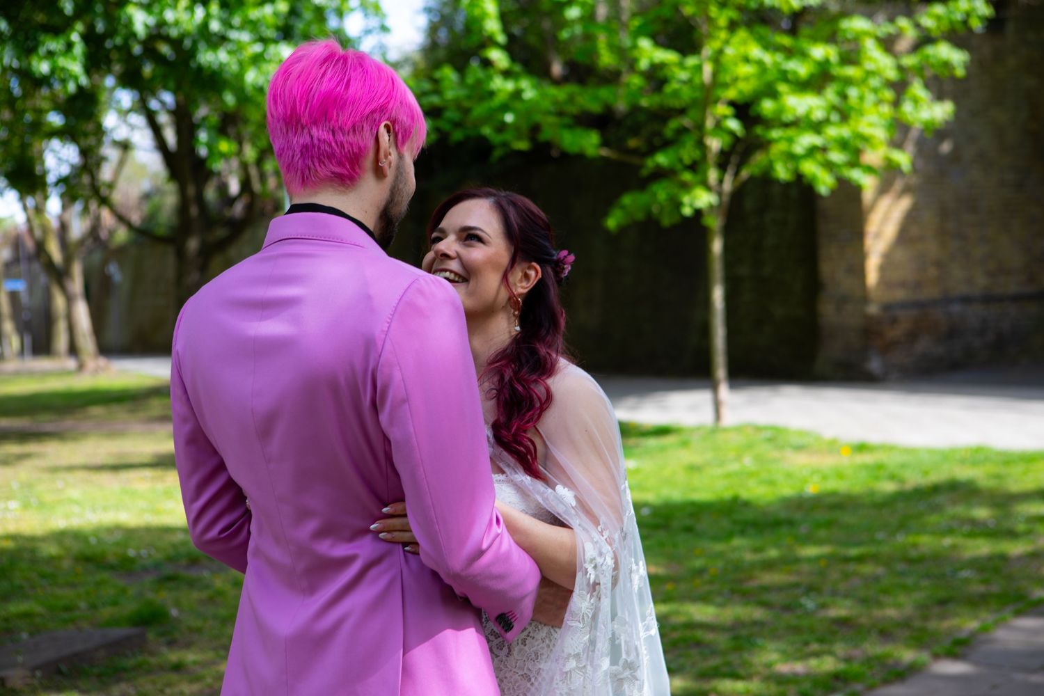 Couple in pink suit and white wedding dress share a romantic moment outdoors in a sunny garden setting.