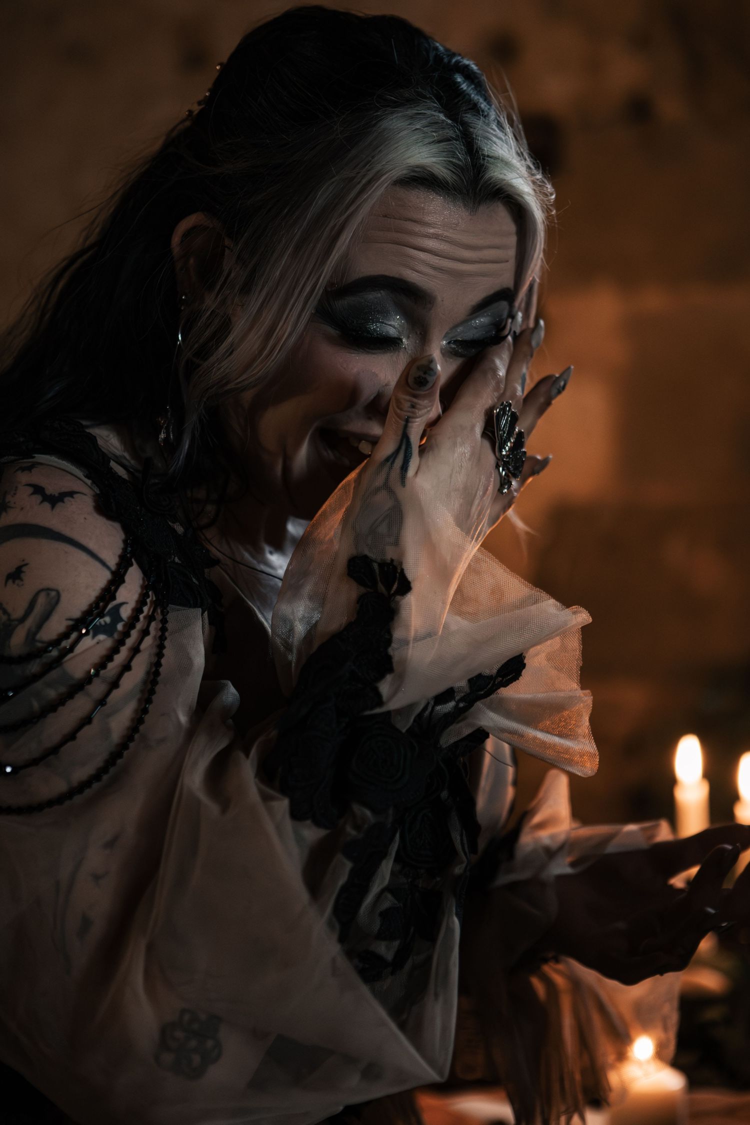 Gothic bride with dramatic black eye makeup and bat tattoos crying by candlelight in ruins.