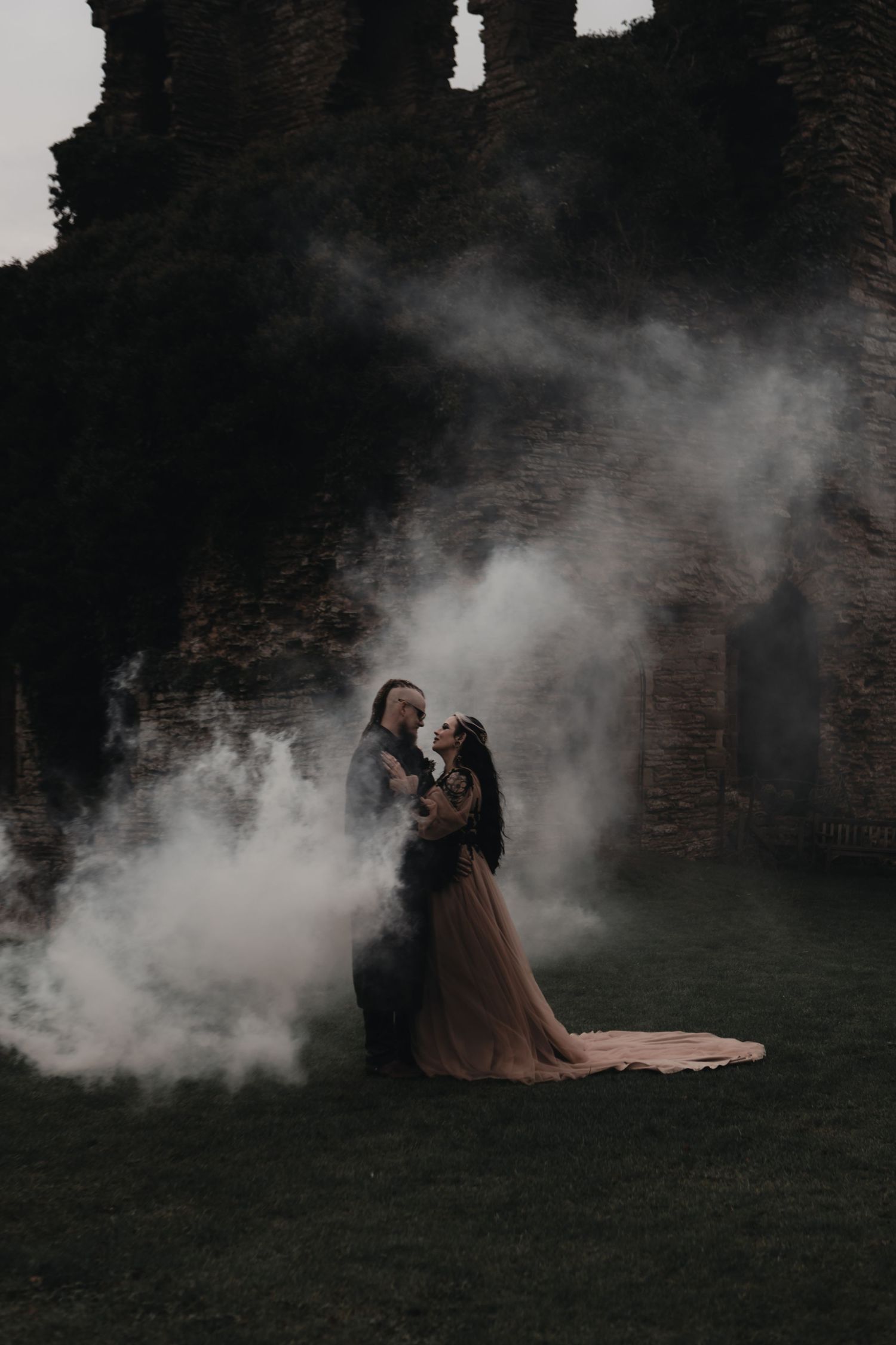 Gothic wedding couple embracing amid dramatic white smoke at ancient stone ruins at dusk.